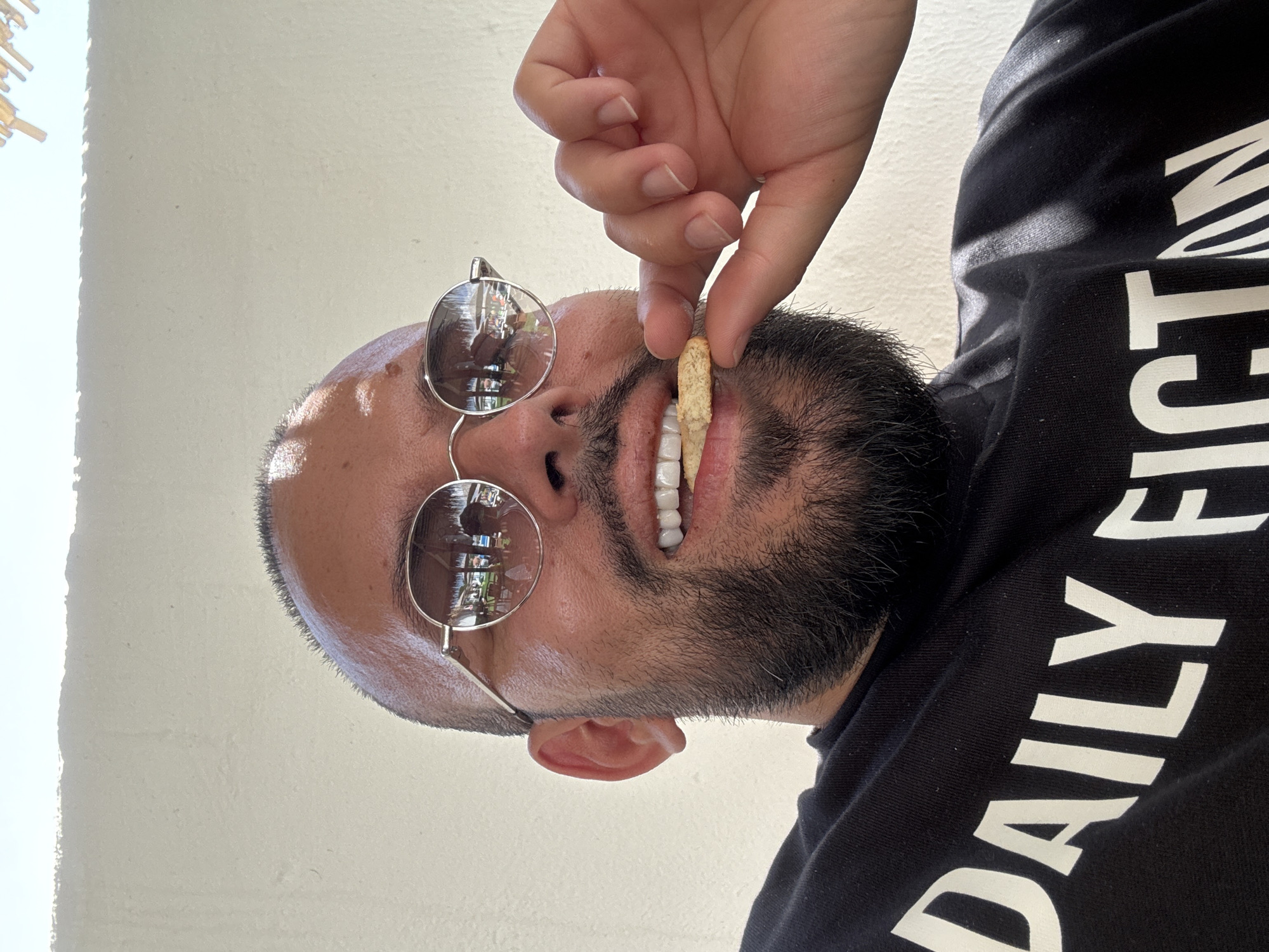 A man is shown from the chest up, looking directly at the camera. He is wearing a black t-shirt with white lettering that reads "DAILY FICTION". The man has a shaved head and a beard, and he is wearing round, silver-rimmed sunglasses. His right hand is raised, and he is holding a biscuit to his mouth, as if about to take a bite. The man's teeth are visible, and they appear to be white and even. The background is a plain white wall, and there is some natural light coming from the top left of the frame. The image appears to have been taken outdoors, possibly in a tropical setting, given the reflections in the man's sunglasses which show palm trees and umbrellas. However, there is no indication of Baliovtsi, Bulgaria in the image.