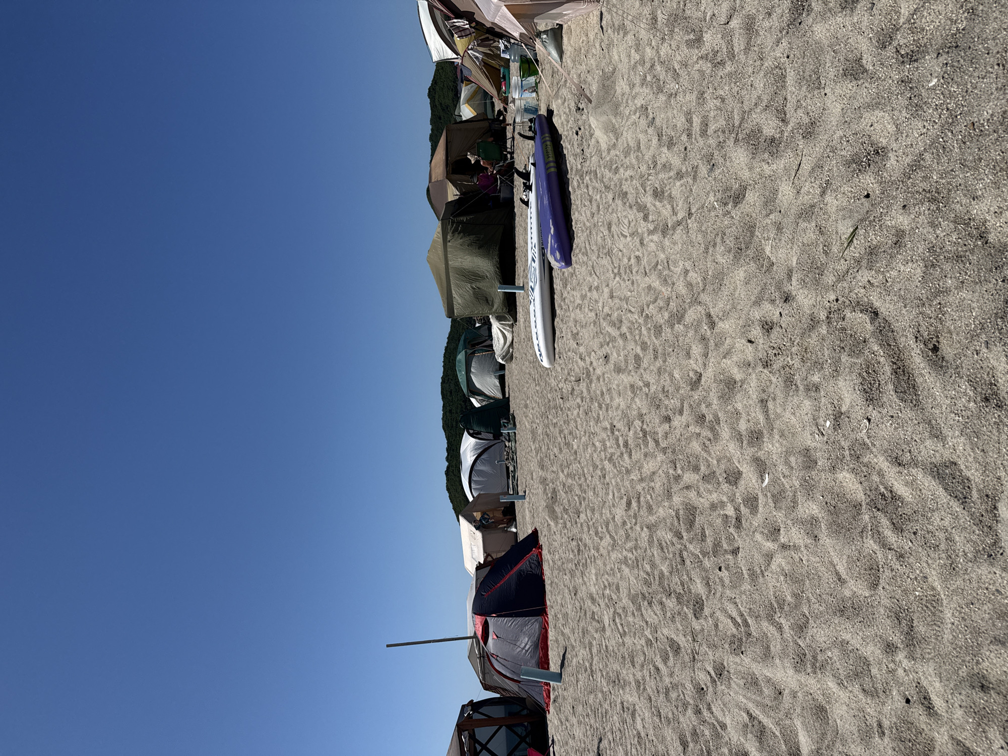 A beach campsite is depicted under a clear blue sky. Numerous tents of various sizes and colors are pitched on the sand, suggesting a recreational gathering. Two surfboards, one white and one purple, are laid out on the sand in the foreground. The environment appears to be a coastal area with trees visible in the distance, possibly indicating the outskirts of Nakhodka, Russia. The scene is tranquil and sparsely populated, with no prominent human activity. The sand is the dominant texture in the foreground, with ripples suggesting the tide may have receded.