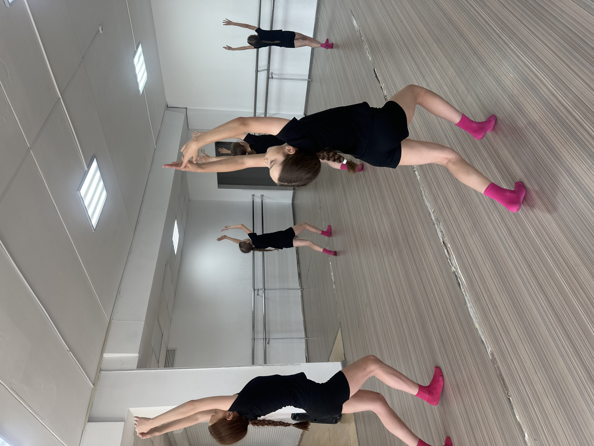 The image depicts a dance studio, likely in Votkinsk, Russia, judging by the architectural style common in the region. There are four young girls visible, all wearing short black dresses and pink socks. They are posed in a dance position, leaning forward with their arms outstretched. One girl is closer to the camera, her hair braided. The studio has a wood-like floor and a barre is visible on the wall. There are mirrored walls, reflecting the dancers and the studio space. The ceiling has recessed fluorescent lights.