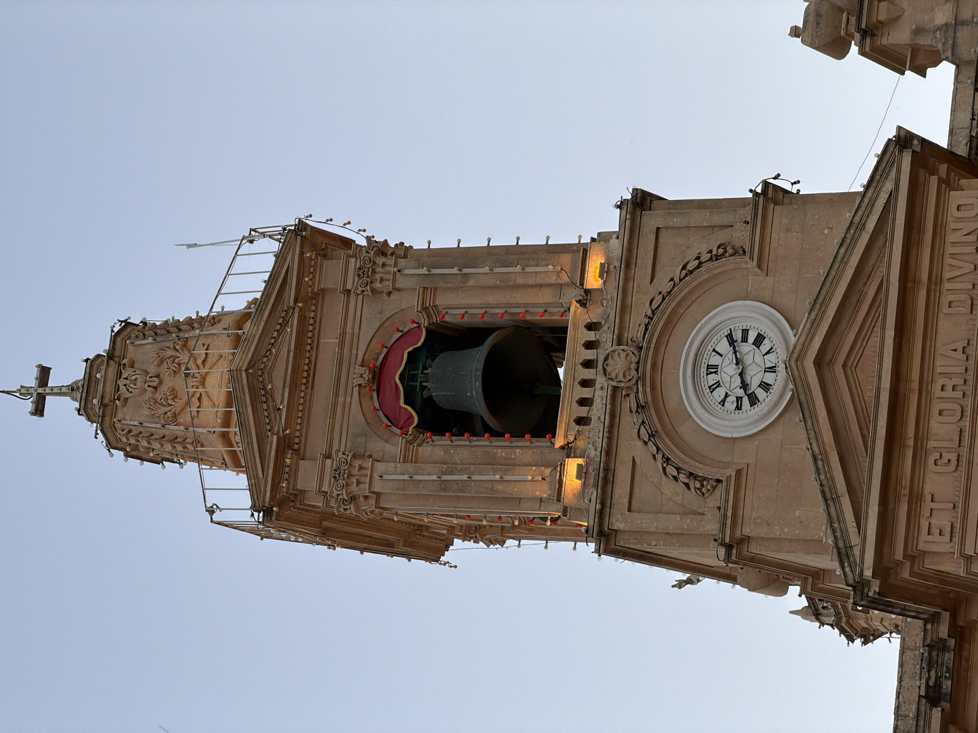 The image shows the upper portion of a historic stone bell tower, likely a church tower, in Fontana, Malta. The tower is made of light brown stone and features ornate architectural details, including columns, carvings, and decorative lights. At the very top, a cross tops the spire, which is adorned with lights and decorative elements. Below the spire, a large bell is visible, partially covered by a red and gold canopy. A clock face with Roman numerals is prominent on a lower section of the tower. String lights are draped around various levels of the structure, some emitting a warm yellow glow, while others appear as small red or white bulbs. The sky is a clear, pale blue, indicating either early morning or late evening light. The inscription "ET GLORIA DIVINO" is visible in large capital letters on a stone ledge near the bottom of the frame. There are no people visible in the image. The scene is static and architectural.