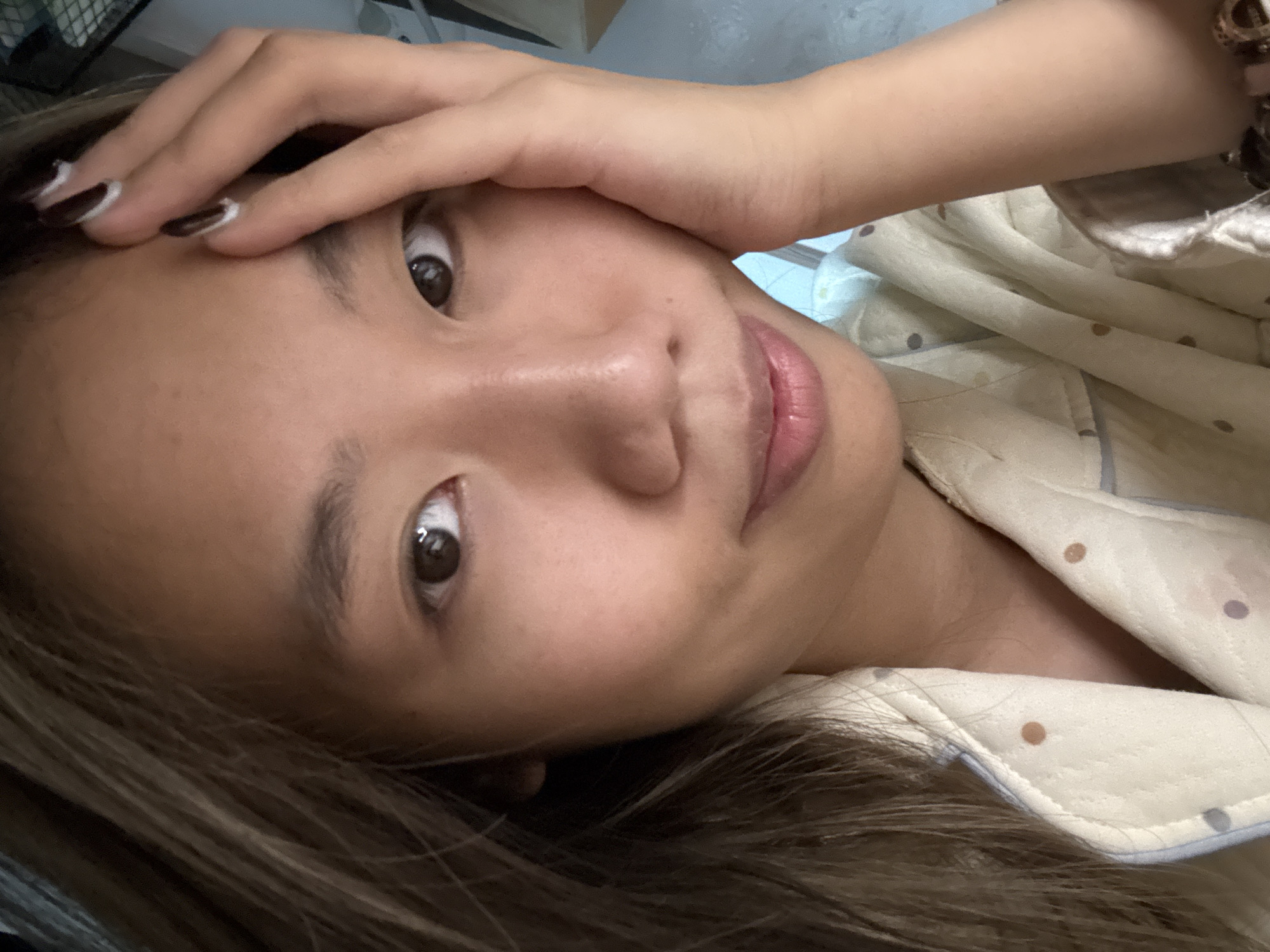 A person with tan skin and brown hair is lying down, looking directly at the camera. The person's right hand is placed on their forehead. The person is wearing a patterned top with brown and blue polka dots. A bracelet is visible on the person's right wrist. The background is blurred. The location is Nan'an District, China.