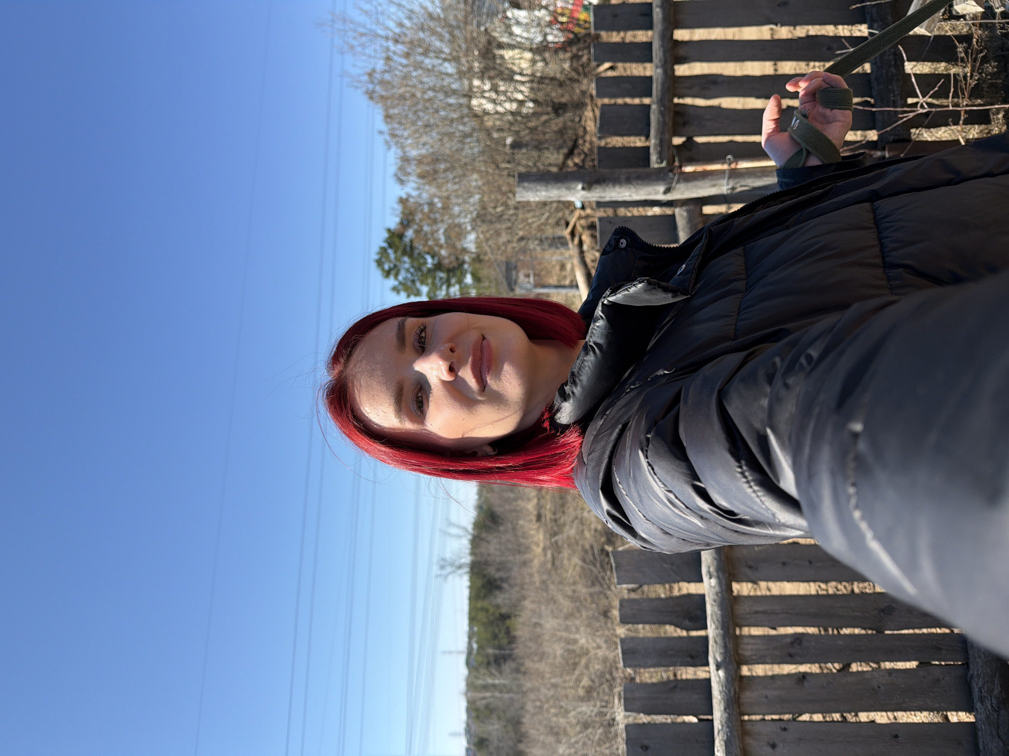 The image shows a woman with red hair wearing a black puffer jacket. She appears to be outdoors in a rural or semi-rural location. A wooden fence or structure is visible to her right. In the background, there are trees and a clear blue sky. Power lines are also visible in the sky. The image provides no information that could identify this location as being in the city of Chaikovsky, Russia.