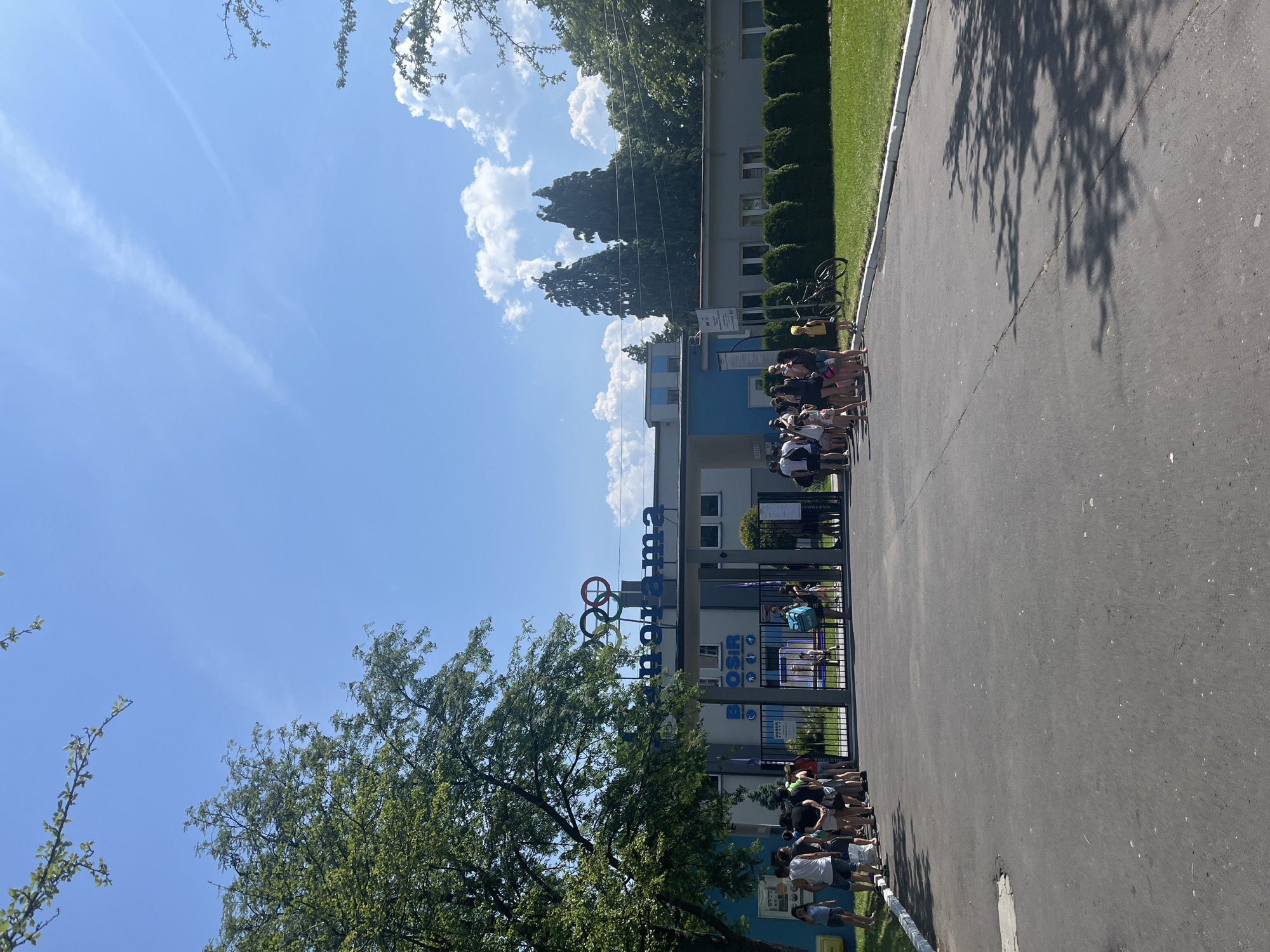 A daytime, eye-level, outdoor shot shows a queue of people waiting to enter a building. The building has a sign with the word "PANORAMA" in large blue letters, and the Olympic rings logo is above it. A banner reads "BOSIR" in blue letters. The people are of various ages and genders, dressed in casual summer clothing. Some are holding bags, and a bicycle is parked nearby. The ground is asphalt, and a green lawn is visible on the right side of the frame. Trees with green leaves are present on the left and in the background. The sky is bright blue with a few white clouds. The scene appears to be a public facility, possibly a sports complex or a recreation center.