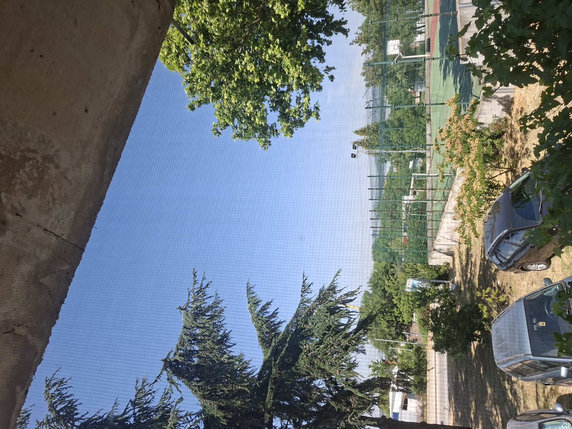 The image captures a view of an outdoor sports court, likely in Aksakovo, Bulgaria, seen through a screen. The sky is a clear, bright blue. In the foreground, a rough, weathered concrete structure juts into the frame from the top, casting a shadow. To the left, the branches of a large coniferous tree with dark green needles hang down. On the right, a deciduous tree with vibrant green leaves is visible.
Below the trees and concrete, an outdoor sports court is discernible. It appears to be a multi-purpose court with a green surface, fenced in by a green metal mesh. A basketball hoop with a white backboard is visible on the court. A concrete wall runs along the front of the court. In front of the wall, there are several bushes and trees with small, light-colored flowers.
In the lower part of the image, two cars are parked on a dry, grassy area. The car on the left is a silver SUV, and the car on the right is a darker colored SUV. The ground is covered in dry grass and some scattered leaves. The overall impression is a sunny day in a semi-urban or suburban setting.