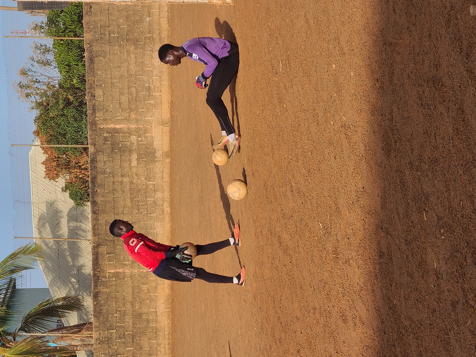The image shows two individuals in athletic attire on a reddish-brown dirt field next to a brick wall. One person is wearing a red long-sleeved shirt and black shorts, holding a soccer ball. The second person is in a purple long-sleeved shirt and black leggings, with two soccer balls between them. A chain-link fence and trees are visible in the background. The photograph was taken outdoors under sunny conditions. The location is suggested to be in the city of Jos, Nigeria.