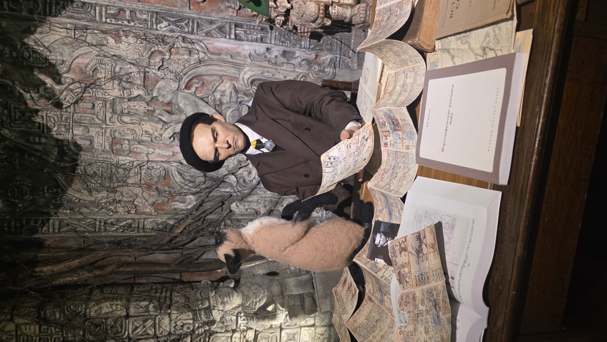 The image shows a diorama display, likely in a museum or exhibit, featuring a wax figure of a man in a brown suit and black hat, seated at a table covered with books and papers. The man is looking at documents with Mayan glyphs. There is a stuffed animal resembling a Siamese cat on the left side of the table. The background depicts carved stone walls, possibly meant to evoke Mayan ruins. The table is a dark wooden color. The scene is set indoors, and the location in the city of Unknown, Unknown is unknown.
