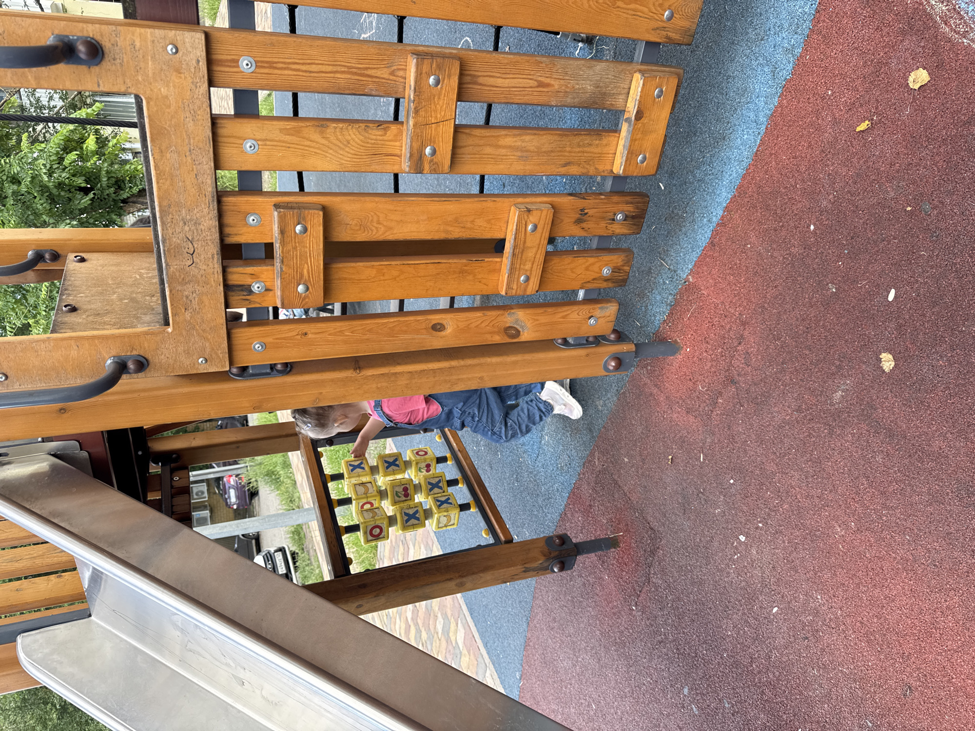 The image shows a young child, likely a toddler, playing a tic-tac-toe game on a playground. The child is wearing blue overalls and a pink shirt, and is positioned on the ground, leaning over the game board. The game board itself consists of yellow cubes with symbols on them, some showing an "X" and others a cherry. The playground appears to be made of wood and metal, with a slide visible in the foreground. The ground is covered with a soft, shock-absorbent material, colored blue and red. In the background, there are glimpses of greenery and some buildings, suggesting an outdoor setting. The time of day appears to be daytime, with natural light illuminating the scene. The overall impression is one of a playful and innocent moment on a playground.