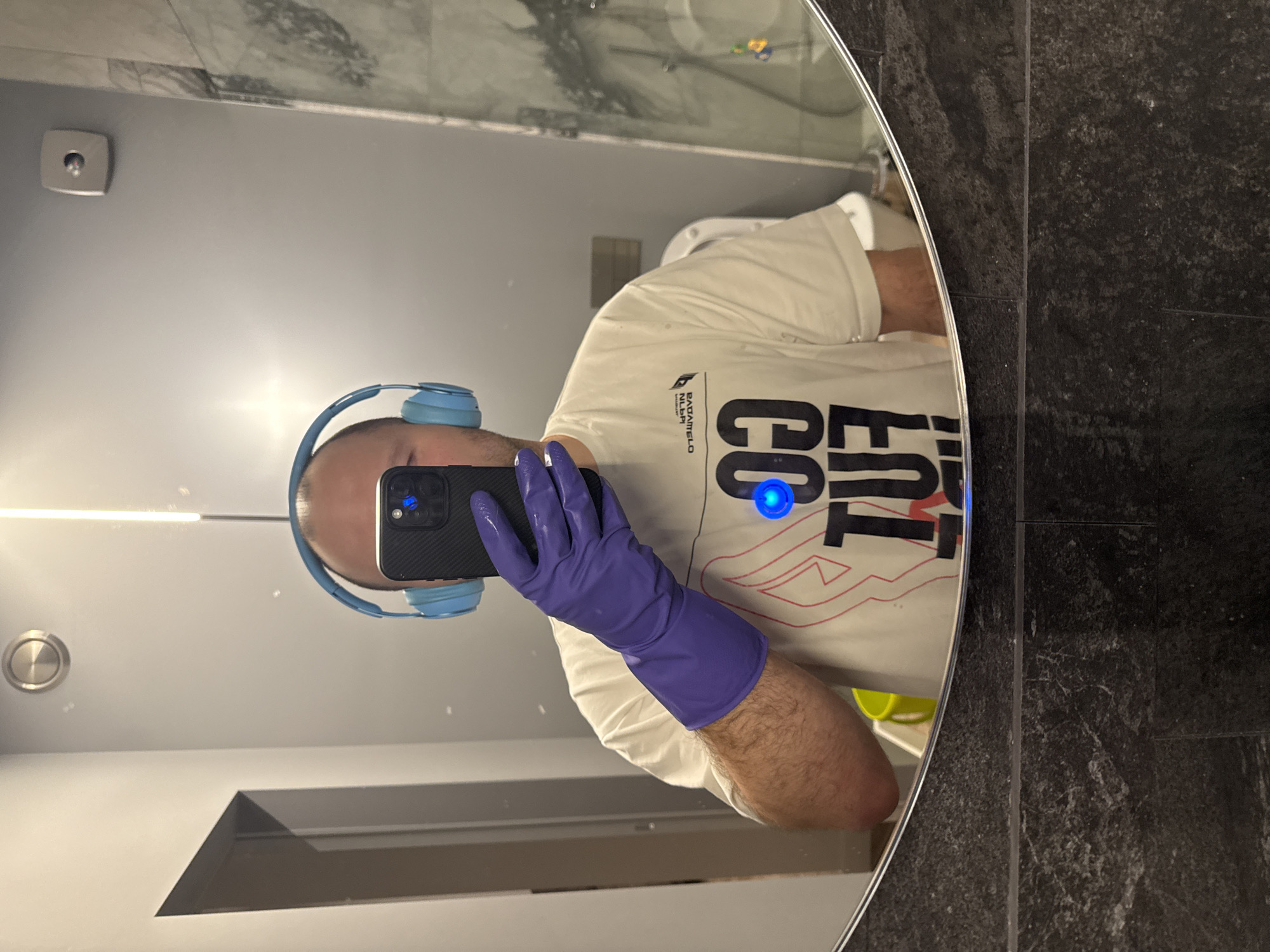 The image shows a reflection in a round mirror. A person wearing light blue headphones and purple gloves is taking a selfie with a black mobile phone. The person is wearing a white t-shirt with black and red lettering. In the background, a toilet and bathroom fixtures are visible. The wall behind the mirror is gray and the wall to the right is dark granite. The city of Krasnoyarsk is not discernible from the image.