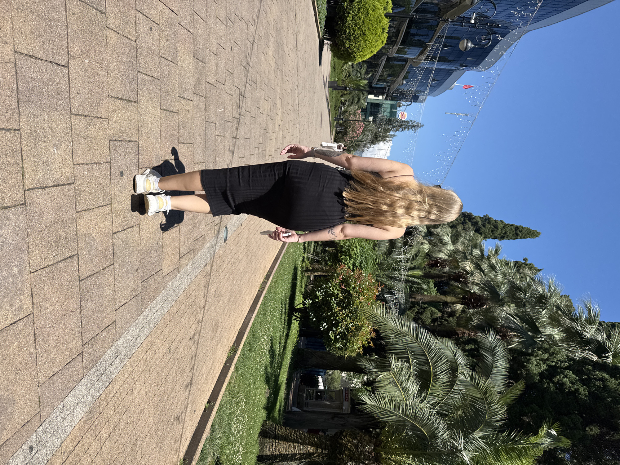 A blonde woman wearing a black midi dress and white sneakers walks on a paved walkway in a park-like setting with palm trees and lush green foliage under a clear blue sky. The walkway is made of tan interlocking pavers with a lighter strip running down the center. To the left, a modern building with a glass facade and decorative lights is visible, along with a neatly trimmed green bush. To the right, a grassy area with small white flowers is bordered by a concrete curb, and several tall palm trees and other dense greenery create a tropical atmosphere. In the distance, another person in white clothing is walking towards the camera. The sun casts shadows on the walkway.