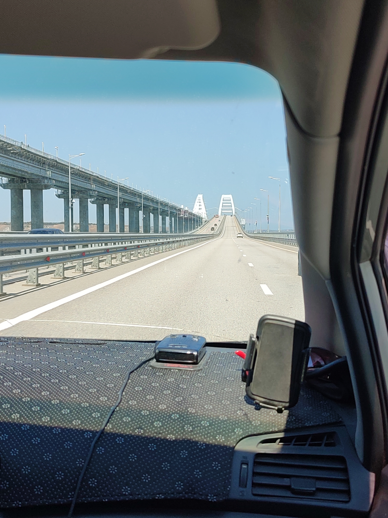 The image is a view from inside a vehicle traveling on a bridge. The road is paved and has white lane markings. On the left side of the road is a metal guardrail, and beyond that are numerous concrete pillars supporting a large elevated structure, likely part of the bridge. On the right side of the road, there is also a guardrail. In the distance, the bridge curves and ascends towards two large arch-like structures. Several vehicles are visible on the bridge ahead.

Inside the vehicle, on the dashboard, there is a radar detector and a phone mount with a black phone in it. A portion of the car's dashboard and a car vent are visible in the foreground. The sky is clear and blue. This scene is likely the Crimean Bridge, which connects Russia to Crimea, and could be near the city of Ilyich, Russia, which is a port city on the Kerch Strait.