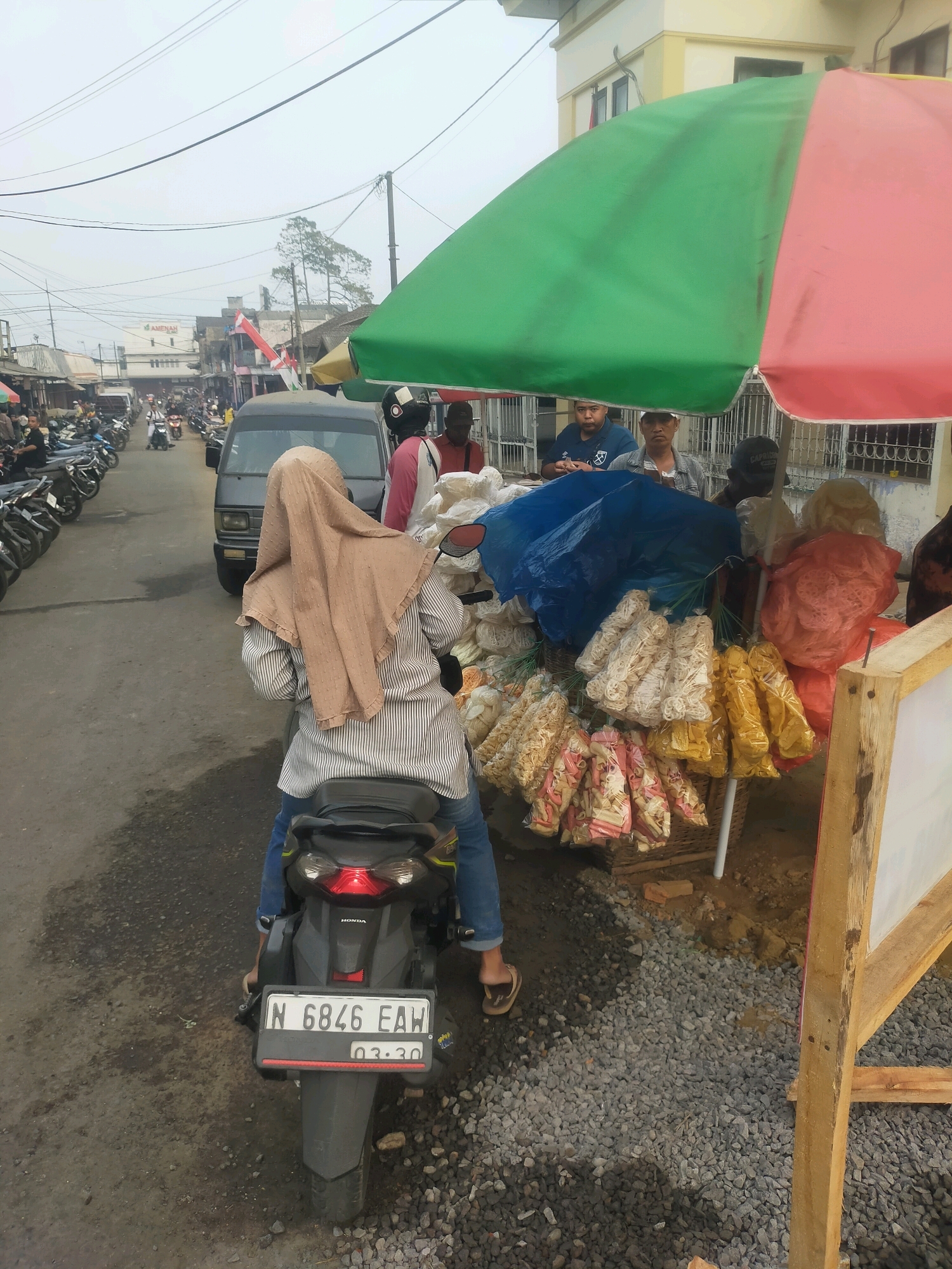In Bululawang, Indonesia, a street scene unfolds with a market. A woman wearing a tan hijab and a striped shirt rides a black Honda motorcycle with the license plate N 6846 EAH, parked on a gravelly street. She is facing away from the camera, towards a stall displaying an assortment of bagged snacks, including some yellow, pink, and white varieties. Behind the stall, under a green and pink umbrella, several individuals are visible, including men. A dark-colored van is parked to the left of the motorcycle. The background shows a row of buildings and parked motorcycles, indicating a bustling marketplace or roadside shopping area under an overcast sky.