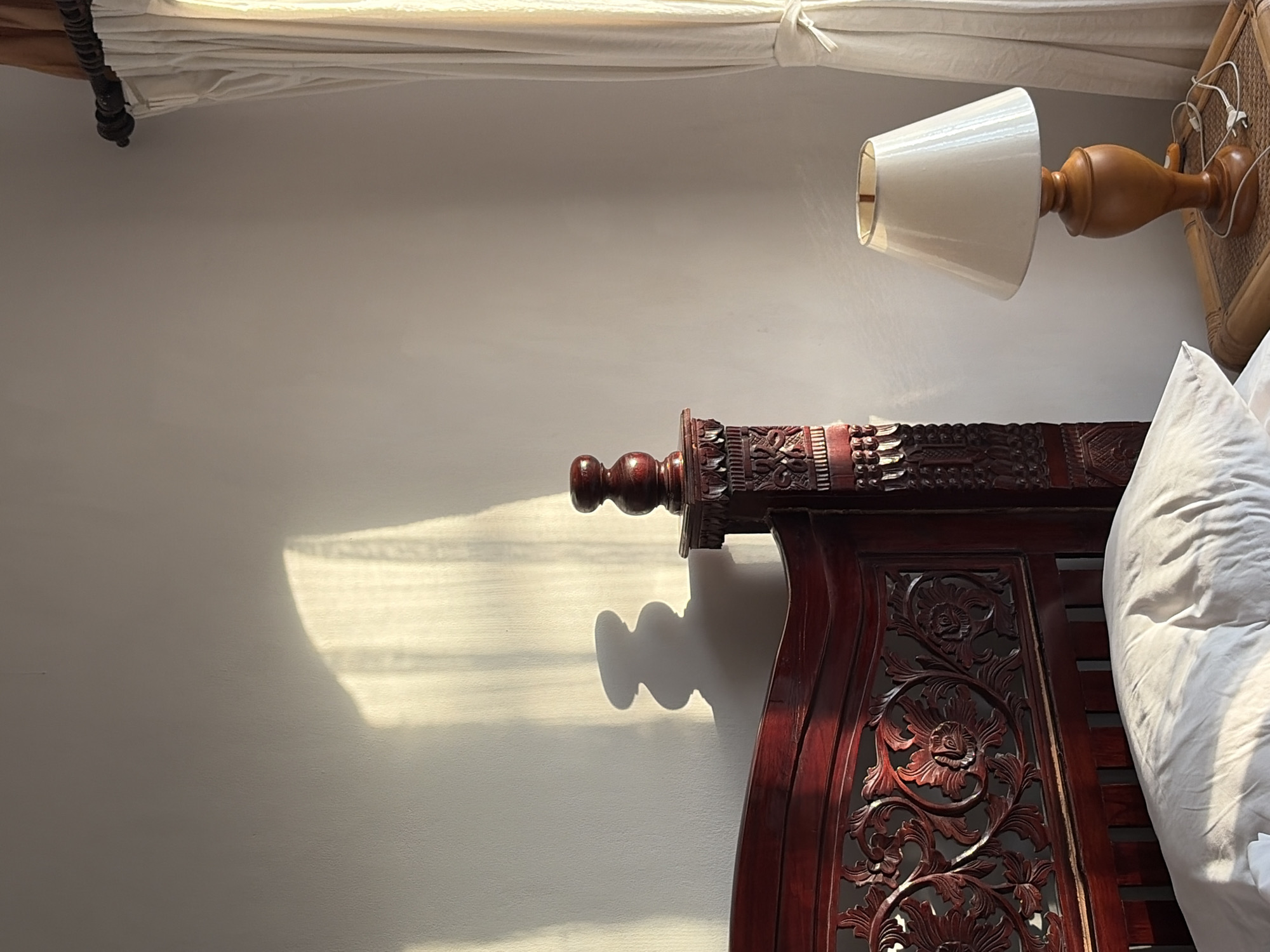 The image shows a bedroom interior in Abang, Indonesia, bathed in the warm glow of natural light. A dark red wooden bed frame with intricate carvings is the dominant feature, with a white pillow resting on it. To the right, a bedside table made of woven material holds a cream-colored lampshade on a wooden base, with its cord plugged into an unseen outlet. A sheer white curtain hangs to the right, partially drawn. A shadow cast by the curtains falls on the wall behind the bed, creating a dappled light effect. The overall scene depicts a serene and cozy bedroom environment.