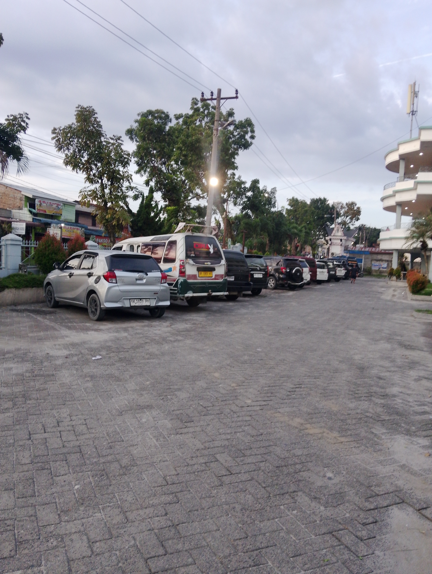 The image depicts a parking lot paved with interlocking concrete blocks in the City of Pematang Siantar, Indonesia. Several vehicles are parked in a row, including a silver hatchback in the foreground with license plate "BK 1819 PY". Behind it is a white van with a green and white front. Further down the line are various SUVs and cars, some of which are dark-colored. Buildings with signs are visible in the background, along with trees and utility poles. The sky is overcast. A person is seen walking in the distance towards the right side of the image.