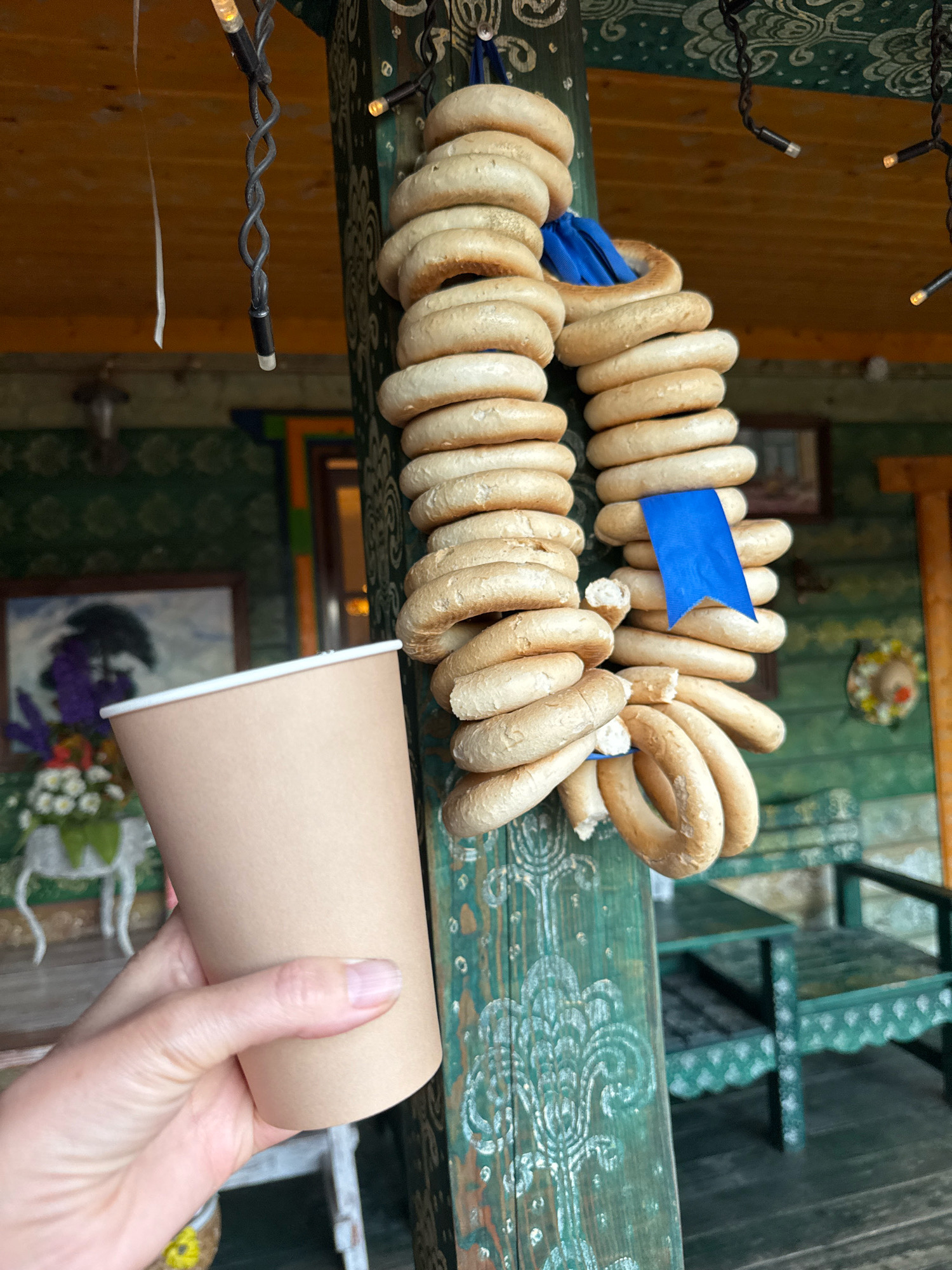 The image captures a close-up view of a hand holding a brown paper cup against a backdrop of a wooden porch and a string of bagels. The porch features green painted pillars with ornate white carvings, a wooden ceiling, and a decorative green awning. String lights with visible bulbs are draped overhead. In the background, a framed painting of a tree and purple flowers is visible on the wall, along with a green wooden bench and chair. A string of bagels, a traditional Russian baked good, hangs prominently from the pillar, adorned with a small blue ribbon. The overall setting suggests an outdoor marketplace or a traditional Russian dwelling in Valday, Russia.