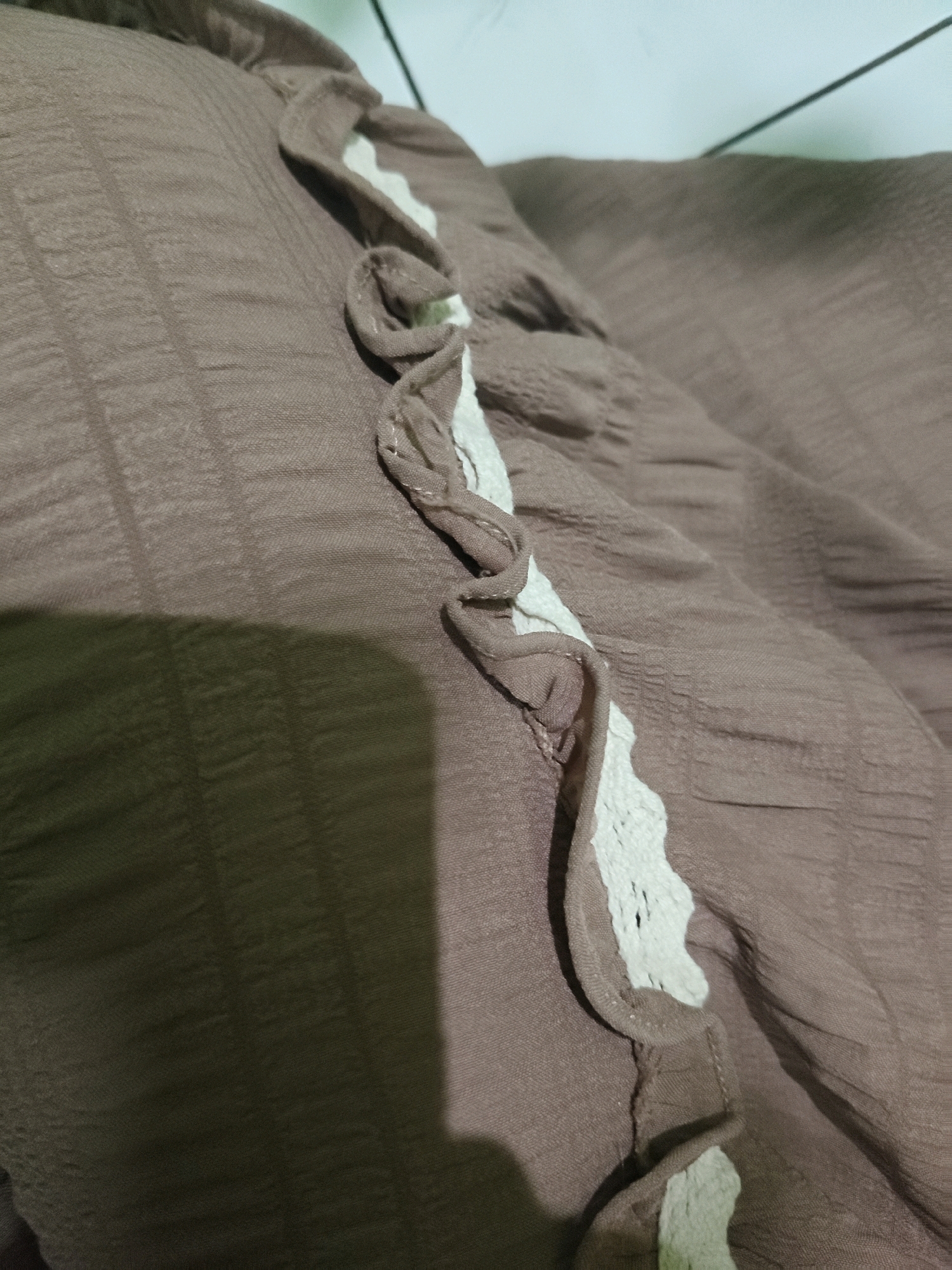 The image shows a close-up, oblique view of a textured, brown fabric with a ruffled, white lace trim running diagonally across it. The fabric has a subtle grid-like pattern. The lighting creates shadows that obscure parts of the fabric, and a dark shadow is cast on the lower left portion of the image. The background appears to be a light-colored surface with a thin black line. The scene is not identifiable as being in Sumberwono, Indonesia.