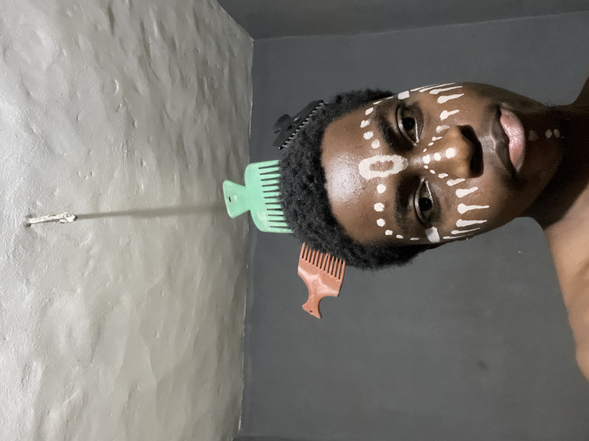 A medium shot depicts a dark-skinned person with white face paint, appearing as dots and lines, oriented sideways. A green hair pick is stuck in the person’s hair, along with a brown hair pick. The person’s body is partially visible on the right, while two gray walls form a corner in the background. A metal rod is affixed to the left wall. It cannot be determined if the image was taken in Nkpor, Nigeria.