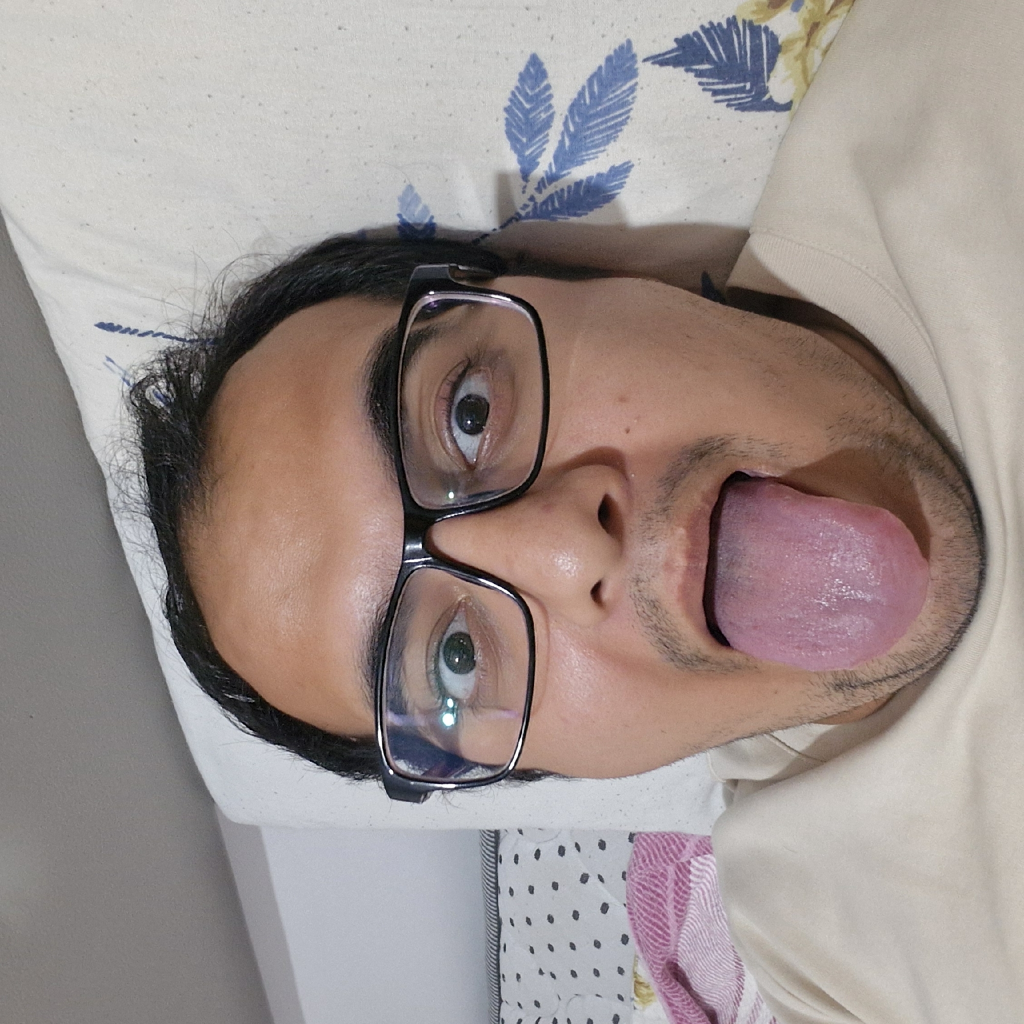 A close-up, forward-facing selfie of a person lying on their back on a bed. The person is wearing black-rimmed glasses and has their mouth open with their tongue extended. The background includes a pillow with a blue floral pattern, a white wall, and a glimpse of a striped pillow and a pink and white striped blanket. There are no discernible landmarks to indicate the specific location within Küçükçekmece, Turkey.