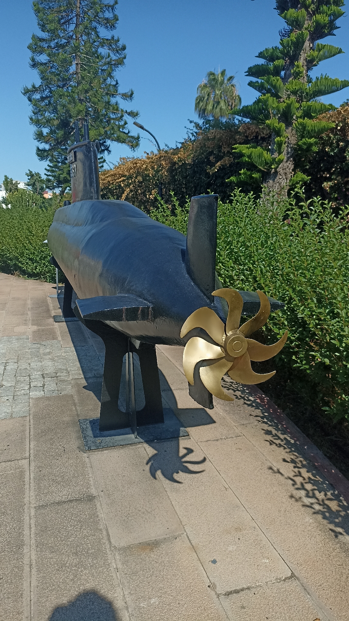 A black, model submarine is displayed outdoors on a paved walkway in Yenişehir, Turkey, under a clear blue sky. The submarine is elevated on black metal stands and has a prominent brass propeller at its stern. Green foliage, including trees and bushes, surrounds the display. Shadows are cast on the ground from the submarine and surrounding vegetation, indicating a sunny day. The scene appears to be a public park or memorial site.