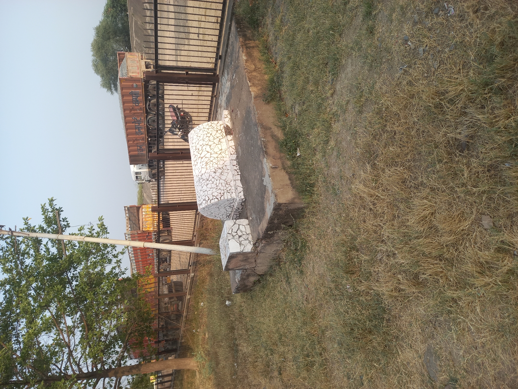 The image shows a roadside scene in Usargaon, India. A concrete structure covered in white mosaic tiles sits on a low platform next to a metal fence. Behind the fence, several trucks are visible. A tree with green leaves is in the foreground on the left. The ground is covered in dry grass.