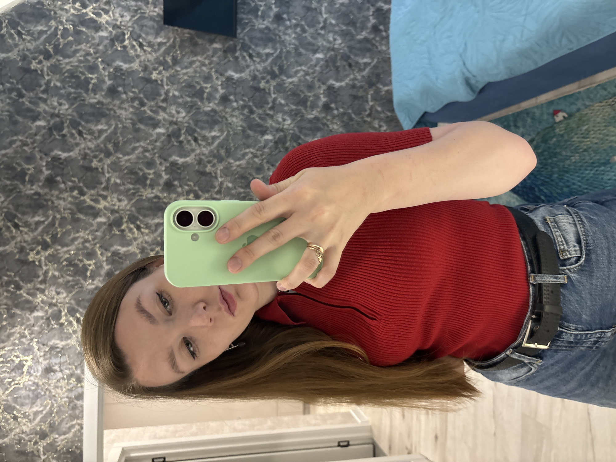 A young woman with long brown hair is taking a mirror selfie in a room. She is wearing a red ribbed shirt with a zipper and blue jeans with a black belt. She is holding a light green smartphone in her right hand. The background shows a dark gray, textured wall with metallic accents, a partially visible doorway with a white frame, and a blue bedspread on the right side. The lighting appears to be indoor artificial lighting. The overall setting suggests a residential interior. The location of Izhevsk, Russia, is not visually verifiable from the image alone.