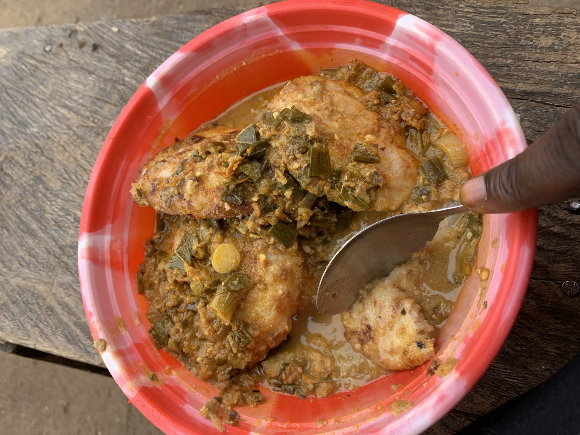 A close-up, overhead shot captures a meal being consumed from a red plastic bowl with white swirled patterns on its rim. A dark-skinned hand is visible on the right, holding a metal spoon that is scooping a portion of the food. The dish features several roundish, light-brown pieces, possibly fried dough, idlis, or fish cakes, generously coated in a thick, greenish-brown stew. The stew is rich with visible chunky ingredients, including chopped green leafy vegetables like scallions and some lighter, disc-shaped vegetable slices. The bowl rests on a weathered, rough-hewn wooden surface, exhibiting prominent grain and wear, indicative of an outdoor or rustic setting. The natural daylight illuminates the scene. The context of Rafin Par, Nigeria suggests this is a local dish being enjoyed. No text is visible in the image.