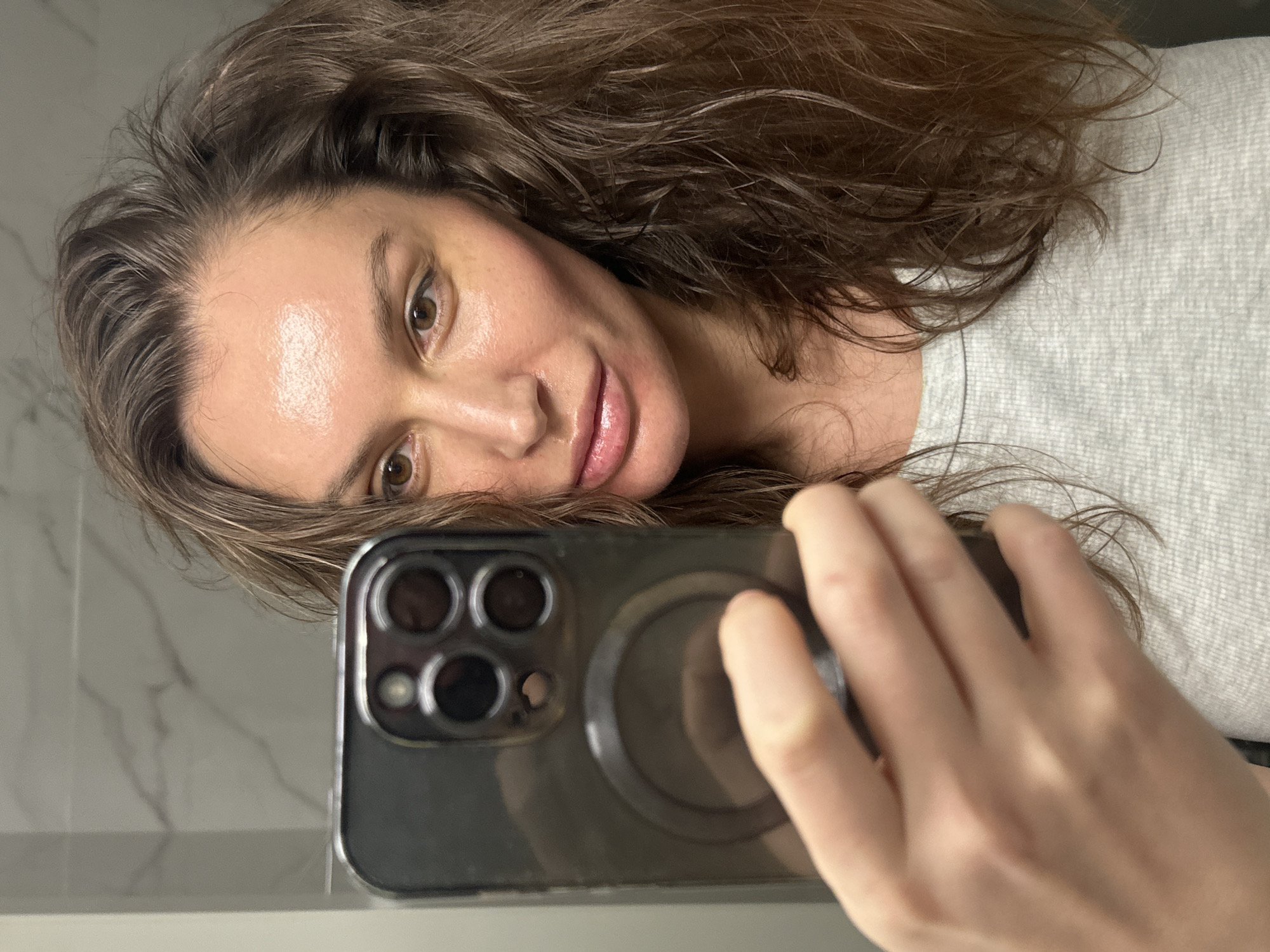 A tilted self-portrait, likely taken in Moscow, Russia, features a woman with long, wavy brown hair, fair skin, and brown eyes. She is captured looking directly into the camera with a relaxed, subtle smile, her lips appearing glossy. She wears a light grey, possibly heathered, casual top, with her hair flowing around her face and over her shoulders.
In the lower right of the frame, the woman holds a dark-colored smartphone in her right hand. The phone is distinctive with its triple-lens camera module and a prominent circular magnetic (MagSafe-compatible) attachment on its back. Her fingers are visible, partially gripping the phone.
The background, visible in the reflection, suggests an indoor setting, likely a bathroom. A light-colored wall with subtle grey marble-like veining can be seen, along with a plain light grey surface, possibly a countertop. The lighting is soft and even, typical of indoor illumination. No discernible text is visible on any subject or object in the image. The overall mood is casual and direct.