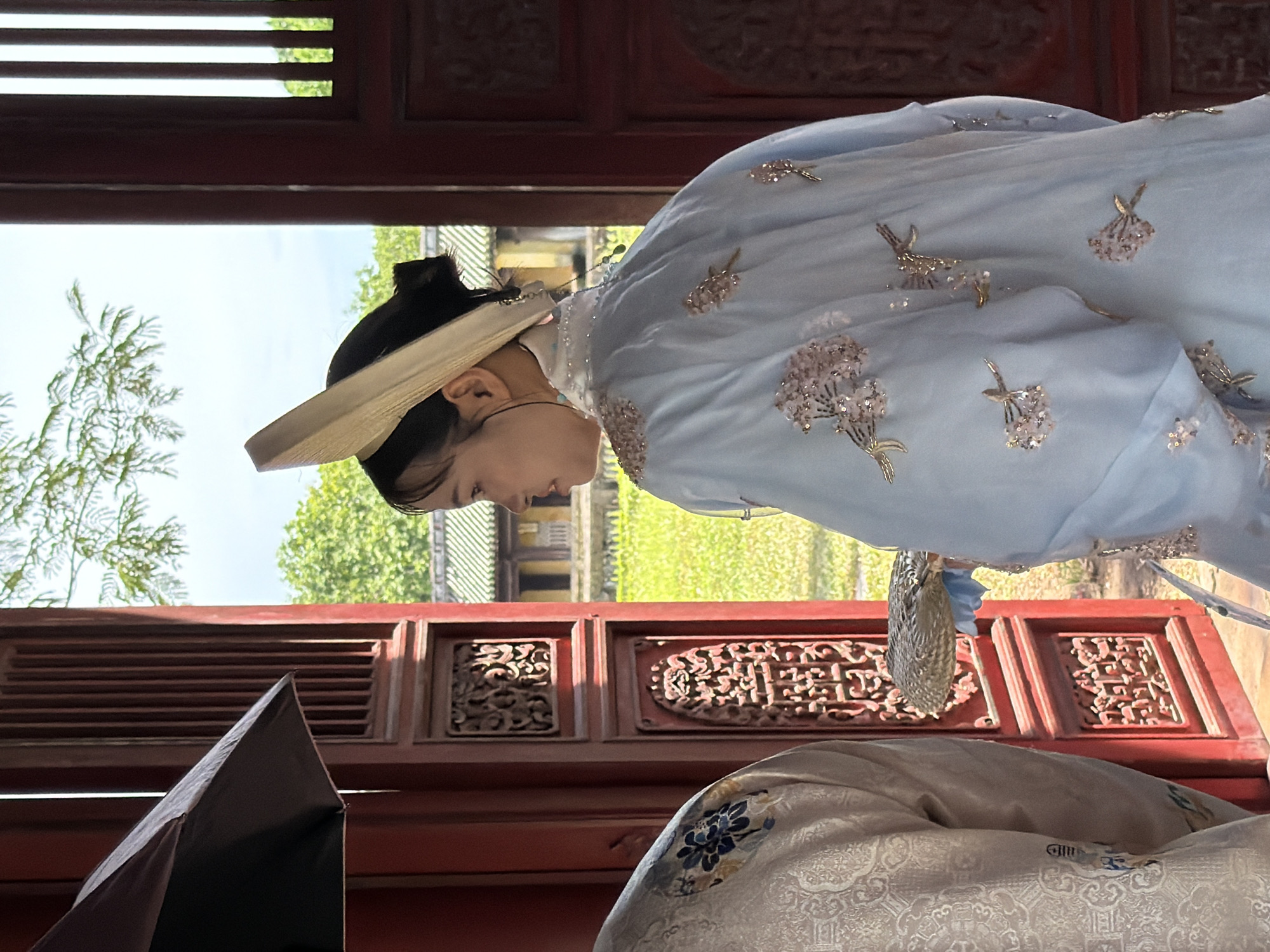 In Phú Xuân Ward, Vietnam, two individuals are partially visible standing near a vibrant red wooden structure. The person on the right, seen from the back, wears a light blue robe adorned with delicate floral embroidery and sequins. They have their hair pulled back and are wearing a wide-brimmed hat, possibly made of straw. In their hand, they hold a woven, fan-like object. To the left, another person's shoulder and upper arm are visible, wearing a cream-colored garment with intricate blue floral patterns. The red wooden structure features ornate carvings and panels, with sunlight filtering through vertical slats, casting shadows and illuminating the scene. Beyond the structure, a grassy courtyard and traditional Vietnamese architecture with green tiled roofs are visible under a bright, overcast sky, with tree branches in the foreground.