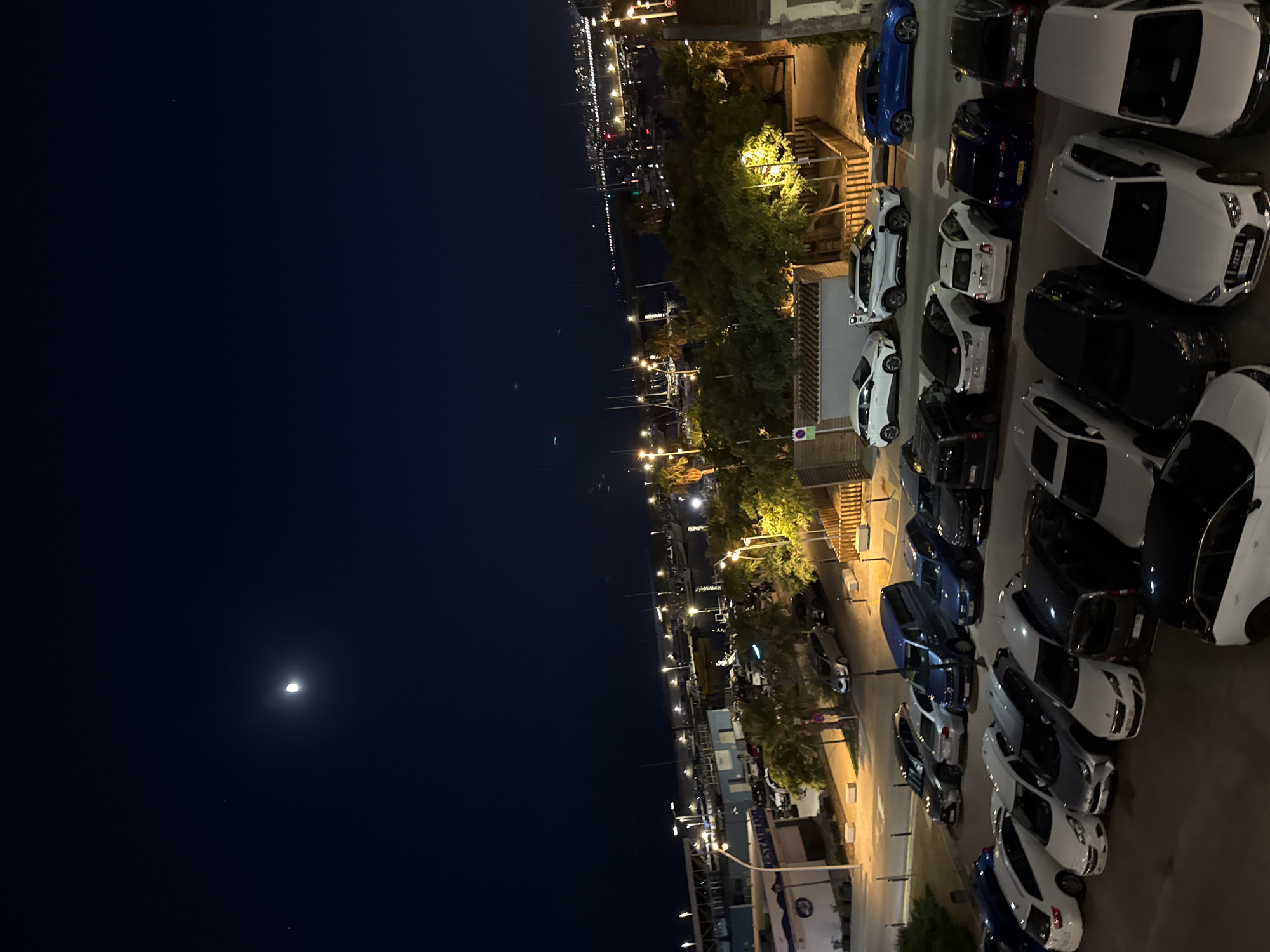 The image captures a nighttime view of a waterfront area, likely a harbor, in Blanes, Spain. A crescent moon is visible in the dark sky, casting a faint glow. The foreground is dominated by a parking lot filled with numerous cars, mostly sedans and SUVs. The vehicles are parked in rows, suggesting an organized parking facility.
Beyond the parking lot, a street lined with trees, some of which are illuminated by streetlights, separates it from a waterfront promenade. Further in the background, a harbor is visible, filled with many sailboats and other vessels. The boats are docked and their masts create a distinct silhouette against the night sky. The lights of the town and possibly other boats create a soft, diffused illumination across the water.
To the left, a building with blue accents and signage that appears to read "RESTAURANT" is partially visible. The overall atmosphere is calm and serene, characteristic of a coastal town at night. There are no visible people in the frame.