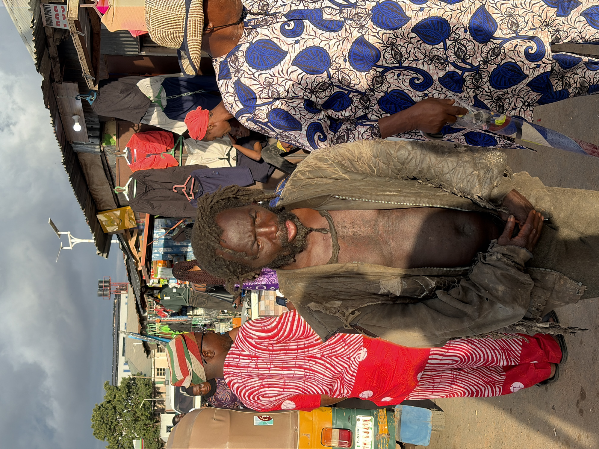 This image depicts a bustling street market scene in Gura Top, Nigeria, characterized by individuals, stalls, and vehicles under an overcast sky with patches of sunlight.
In the immediate foreground and center stands a man with dark skin, dreadlocked hair, and a full beard. He wears a tattered, dark olive-green zippered jacket, unzipped to reveal his bare chest and abdomen, and dark trousers. His hands are loosely clasped in front of him, and his mouth is slightly ajar with a somber expression.
To the left of the central figure, with his back partially turned to the viewer, is a man wearing a vibrant red long-sleeved tunic and matching trousers, both adorned with a white patterned design of concentric circles and lines. He wears a multi-striped cap with red, white, and green bands. He appears to be holding a dark object, possibly a phone, to his right ear. Immediately behind him, to the far left, is the rear of a yellow and green auto-rickshaw, commonly known as a Keke Napep. A white license plate, "PLATEAU YM-645AA," is visible on the vehicle. A woman in a patterned purple garment is partially visible behind the man in red.
To the right of the central figure, a man in a patterned blue, white, and black long-sleeved tunic stands facing slightly left. He wears a light-colored, possibly straw, hat that partially obscures his face. His right hand holds a long, translucent plastic-wrapped item, possibly a rolled-up poster, umbrella, or toy, featuring colorful patterns.
The background consists of numerous market stalls constructed with corrugated metal roofs and rough wooden supports. Various items of clothing, including jackets, shirts, and trousers in different colors, are hung for display and sale on hangers. A white light bulb is visible under one of the stall roofs. A partially visible sign above a stall reads "MONI POINT DCC." Further in the background, a tall pole with solar panels and lights is visible, along with a red tower supporting a water tank. Several multi-story buildings are faintly visible in the far distance under the cloudy sky. The ground appears to be an unpaved or dirt surface.
