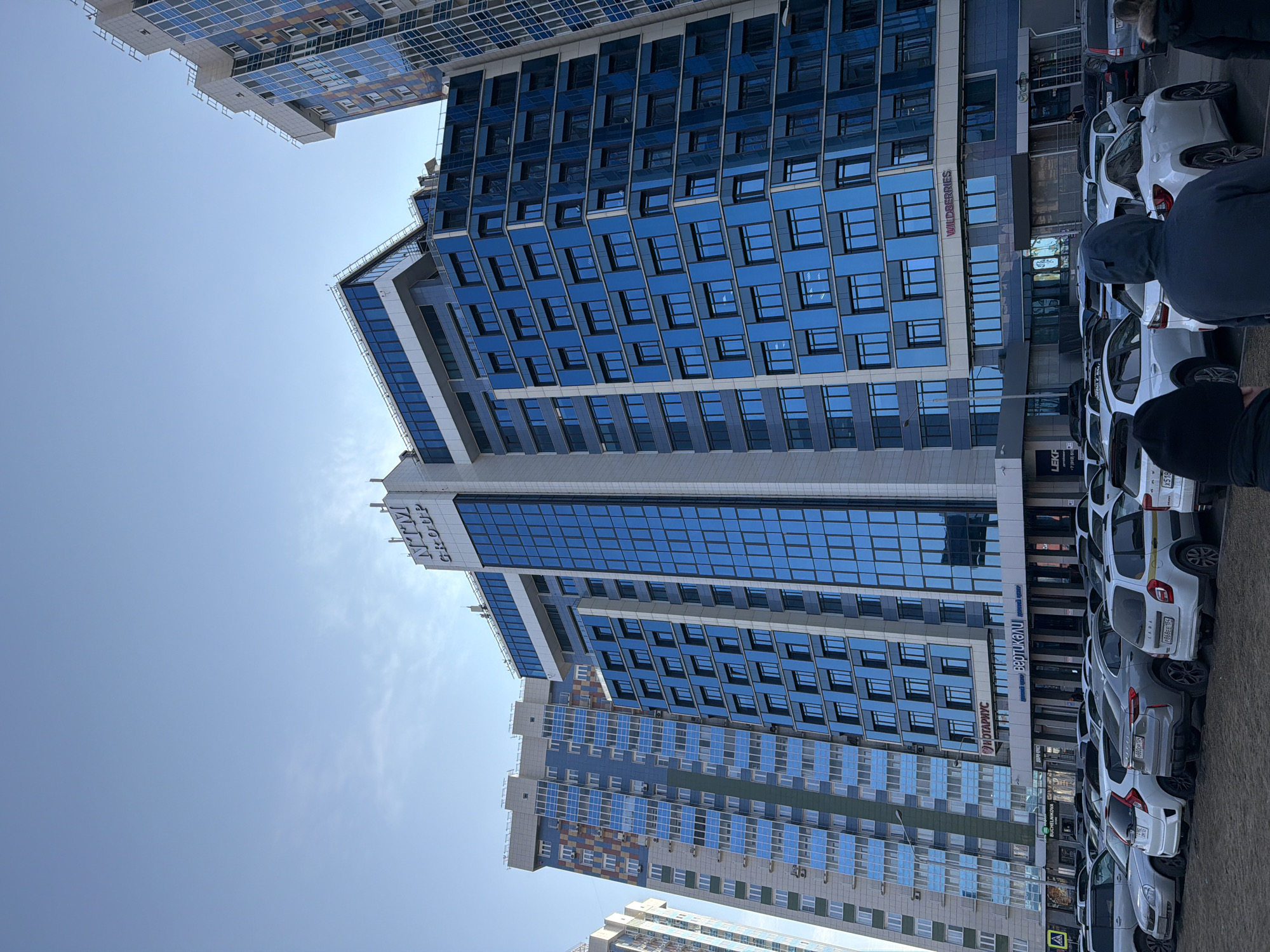 The image depicts a multi-story building with blue glass windows in Krasnoyarsk, Russia. The building has a sign that reads "NTM Group" in Cyrillic script. Adjacent to the building is a row of parked white cars. Some people are present in the foreground, partially obscured. The sky is clear and blue. Another building is visible in the background. A sign for "Wildberries" in Cyrillic script is visible lower on the building.