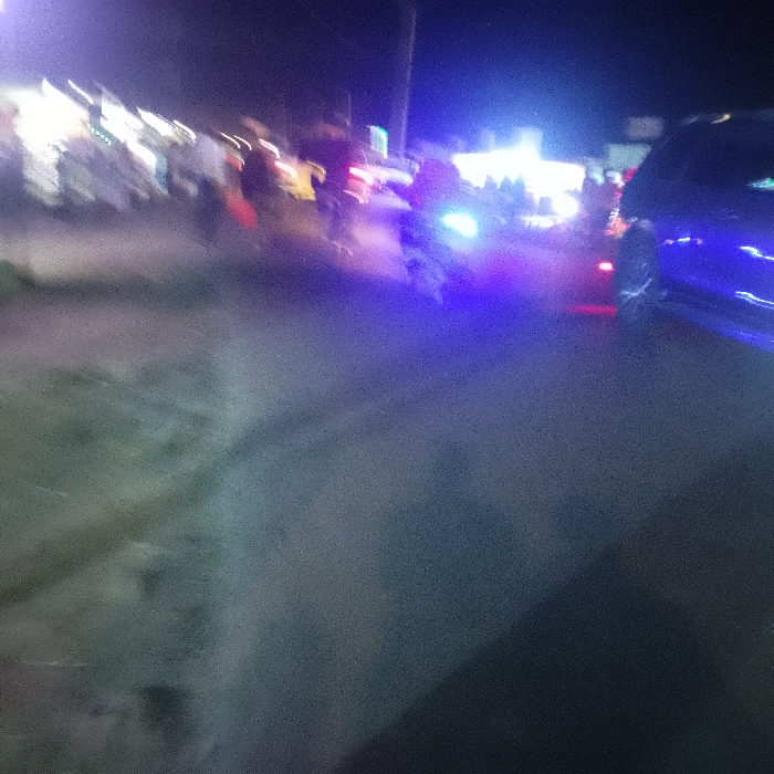 A blurry, grainy night scene shows a street with people gathered. A bright blue light emanates from the center, possibly from a vehicle or street lamp. Several figures are illuminated by the light, though their features are indistinct. A blue car is partially visible on the right edge of the image. The road is dark and blurry, leading toward the bright light source.