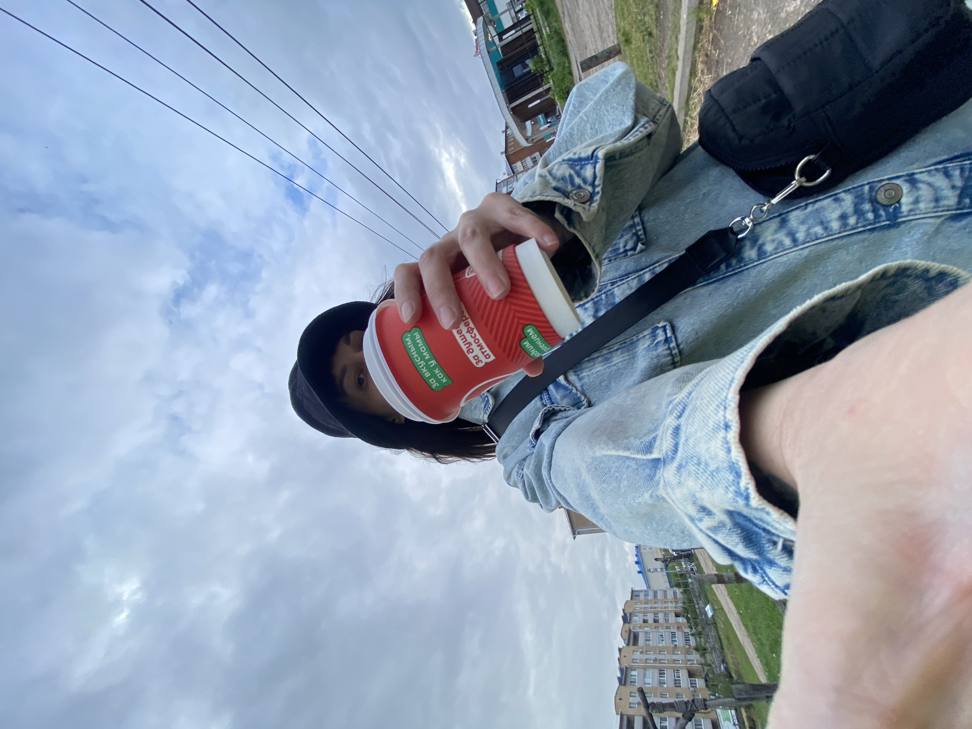 A low-angle photograph depicts an individual holding a red paper cup with Cyrillic text, possibly containing a beverage, in front of an urban landscape. The person wears a light blue denim jacket, a black hat, and a black cross-body bag. Electrical power lines stretch across the upper portion of the frame against a cloudy sky. In the background, a few buildings are visible, including a light-colored apartment building with multiple windows. The buildings are consistent with architecture found in Votkinsk, Russia. The photograph likely taken during daytime.