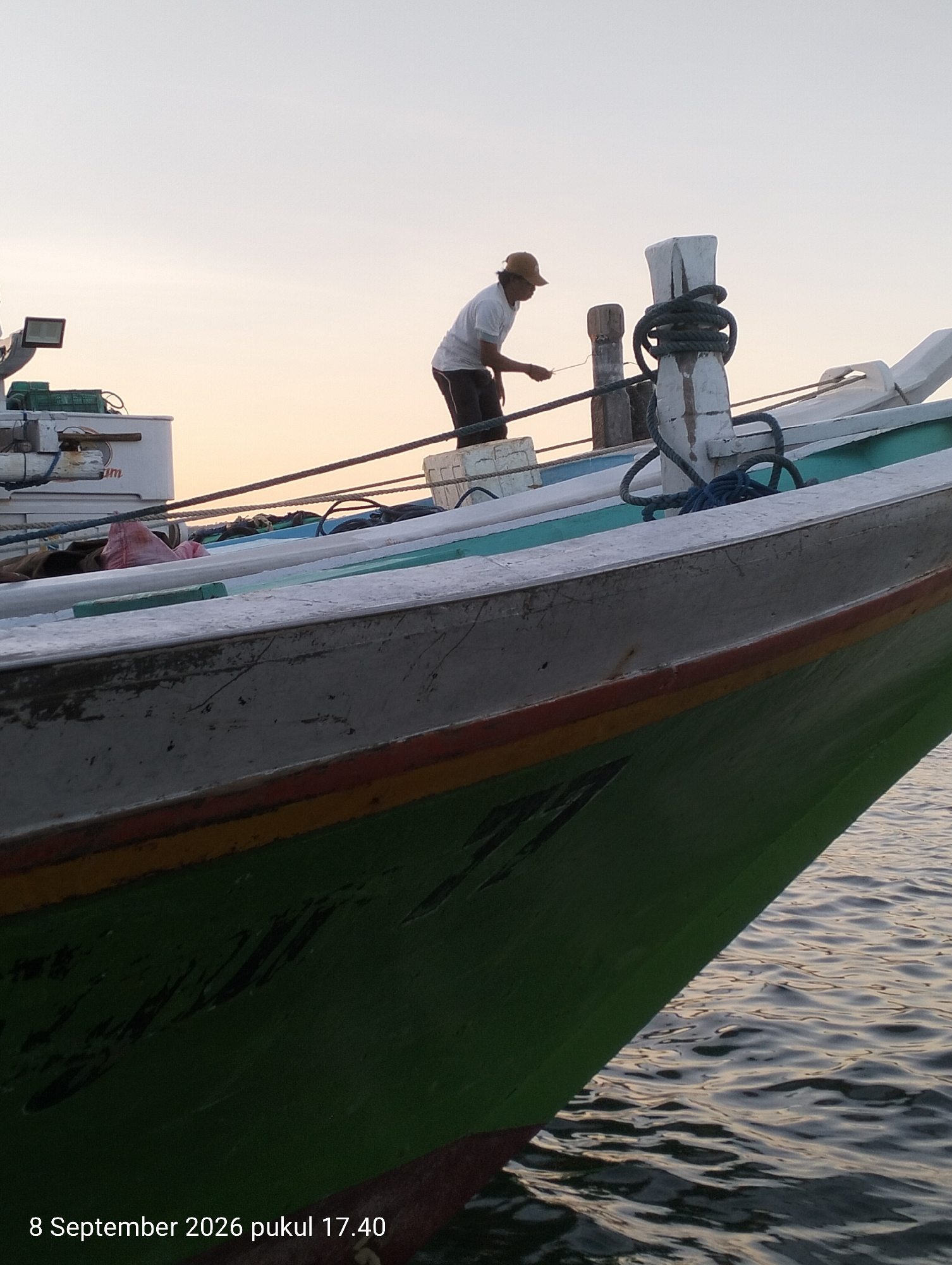 The image shows a fishing boat docked or anchored in the water. A lone fisherman, wearing a white t-shirt and a tan cap, is standing on the deck of the boat. He appears to be tending to ropes or fishing gear. The boat has a green hull with white and blue accents. In the background, there are other boats and structures, suggesting a harbor or a busy waterfront. The sky is overcast with soft, warm light, indicating either sunrise or sunset. The water is calm with gentle ripples. A date stamp in the lower-left corner reads "8 September 2026 pukul 17.40", which translates to "September 8, 2026 at 17:40". This timestamp and the style of the boat suggest the location is likely Kendari, Indonesia, a coastal city known for its fishing activities and traditional boats.