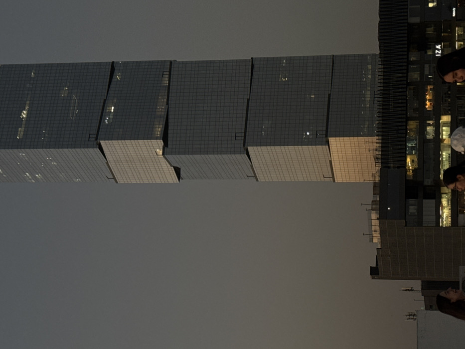 The image shows two modern high-rise buildings at dusk or night. One building has a unique staggered or tiered design, with sections that appear to jut out slightly from the main structure. Its facade is made up of many small windows creating a grid pattern. The second building is more traditional and also has many windows that are lit up in some sections. There are a few people visible in the bottom right of the frame. The sky is gray.