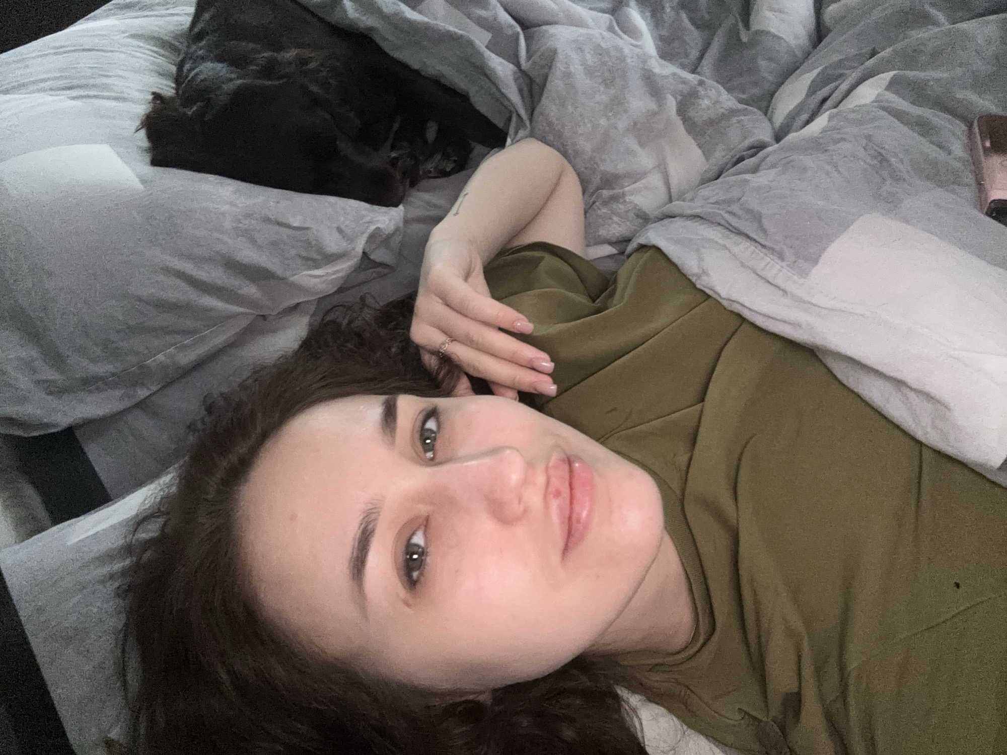 A woman is lying in bed with a dark-colored dog, presumably in a residential location in Марковский, Russia. The woman is wearing an olive-green shirt and has dark, curly hair. She is looking directly at the camera. The bedding is gray and white. A phone is visible near the upper right corner of the frame. The focus appears to be on the woman's face.