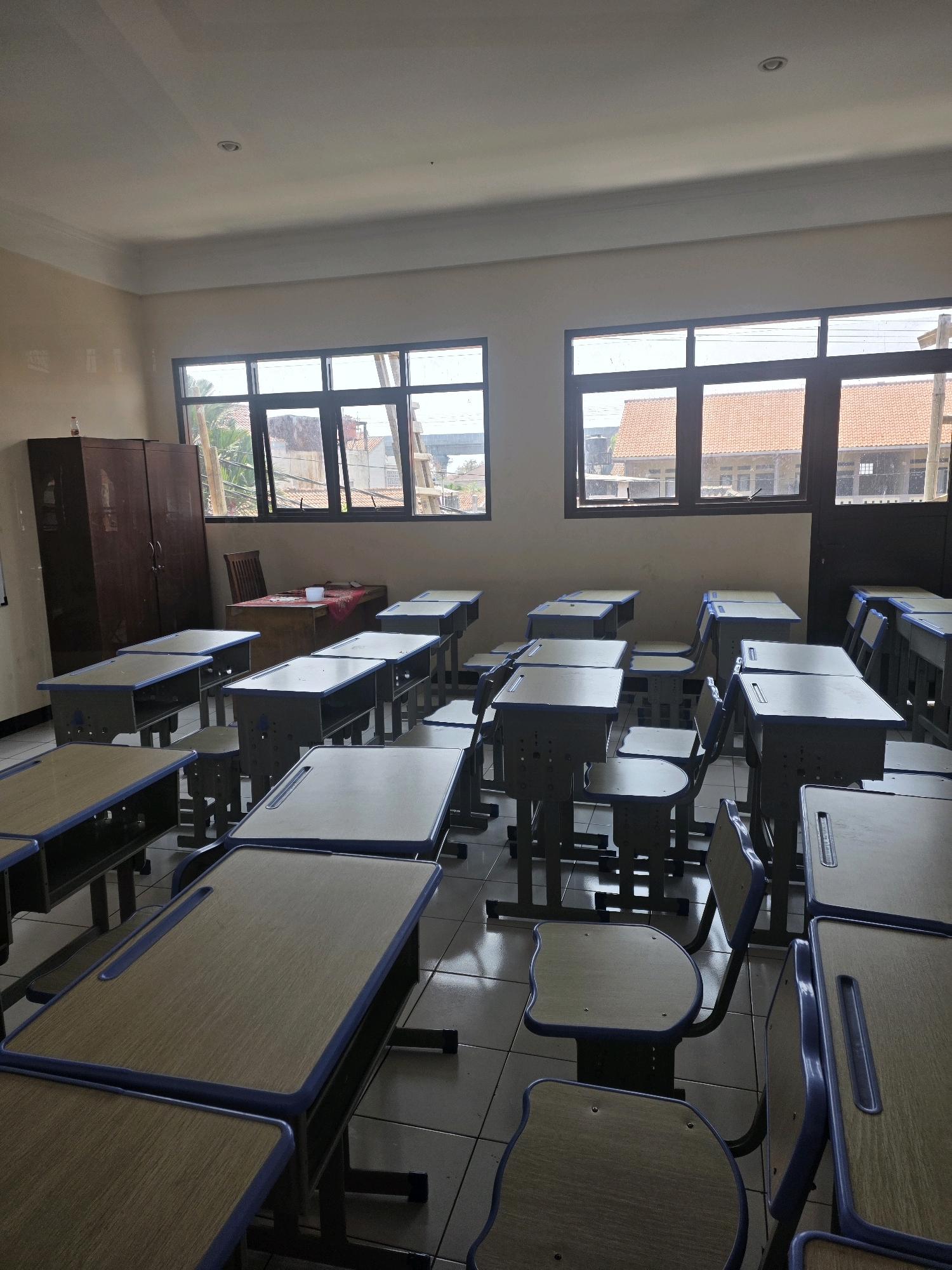 The image depicts an empty classroom in Bojongsoang, Indonesia. The room is furnished with numerous identical wooden desks and chairs, arranged in rows. The desks are light brown with blue trim, and the chairs have blue seats and backs. A large wooden cabinet stands against the left wall. Two windows with dark frames overlook buildings with orange tiled roofs. Sunlight streams through the windows, casting shadows on the tiled floor. A teacher's desk is situated near the windows, with a red cloth and a white cup on its surface. The overall impression is of a well-maintained, but currently unoccupied, educational space.