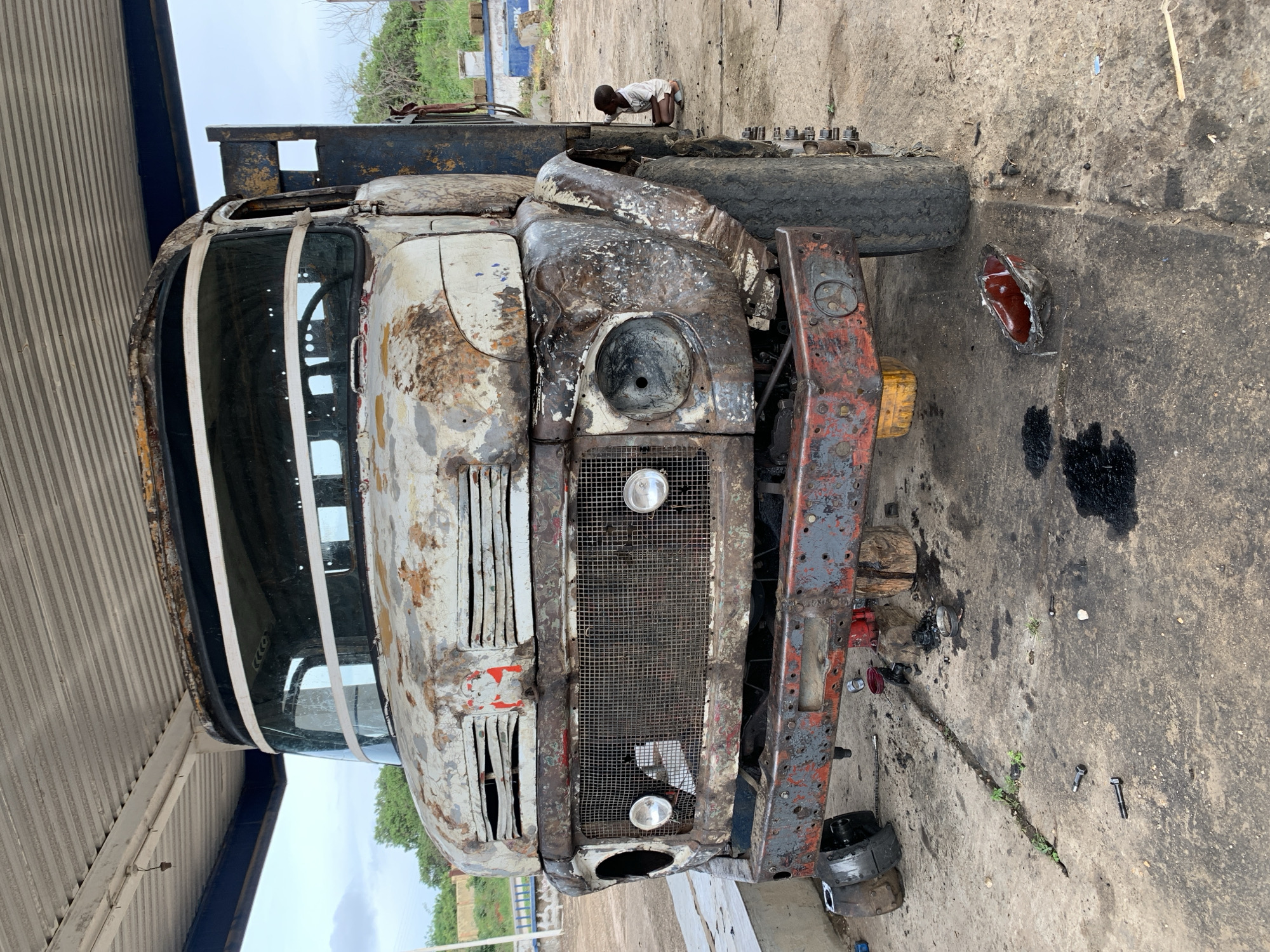 The image shows the front of a heavily rusted and damaged truck, possibly undergoing repair, under the partial shelter of a roof structure, likely a garage or workshop. The truck's front end is heavily corroded, with missing headlight lenses and deteriorated body panels. The grill is intact. Various parts and tools are scattered on the oily concrete floor around the truck. A child is atop the truck, appearing to interact with it, while the background reveals an outdoor environment. Based on the context, the image is likely taken in Gbeleko, Nigeria.