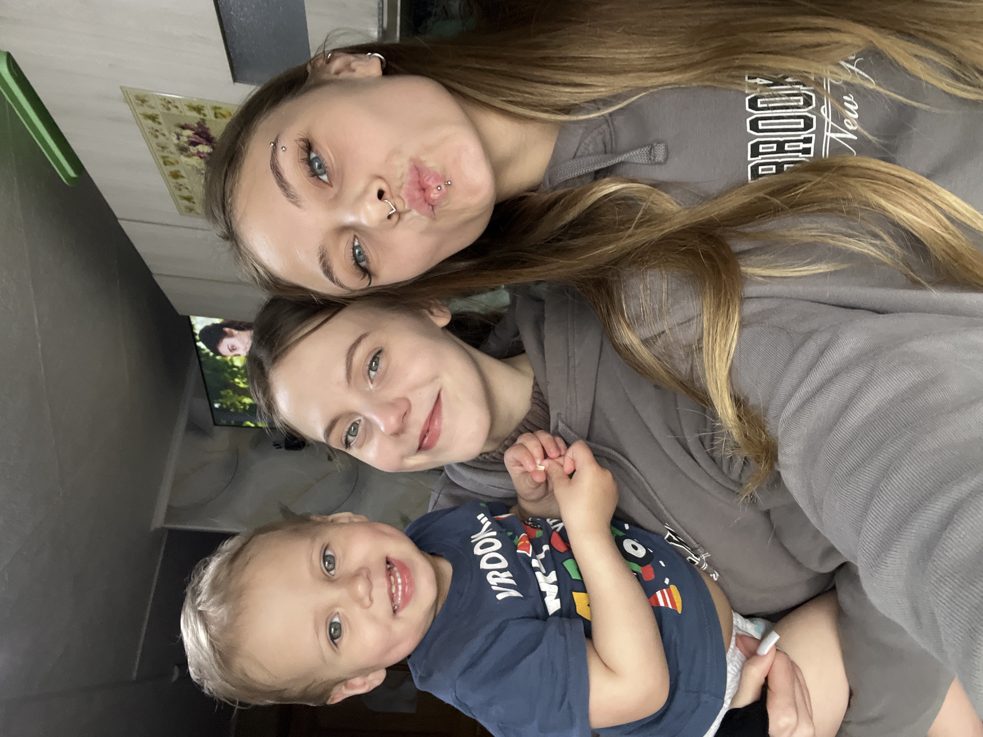 An indoor, casual group portrait features three individuals: two young women and a toddler, captured in a selfie-style photograph.
The uppermost woman has long, light brown hair, blue eyes, and multiple facial piercings, including an eyebrow piercing, a septum piercing, and a labret piercing below her lower lip. She is wearing a grey hoodie with "BROOKLYN New York" visible text and is playfully pouting for the camera.
Below her is another young woman with light brown hair and light-colored eyes, smiling warmly. She is also wearing a grey hoodie. Her hand, with light-colored manicured nails, is visible near the toddler.
The toddler, positioned at the bottom, has light hair and blue-green eyes, displaying a wide, cheerful smile. The child is wearing a dark blue t-shirt with colorful graphics and the word "BROOKLYN" visible, along with a diaper.
The background shows a simple home setting with a wall decorated with a framed picture or tapestry featuring floral patterns. A television screen is partially visible in the background, displaying a person. A green object can be seen on the left side of the background. The overall mood of the image is joyful and relaxed, highlighting a familial or close relationship between the subjects.