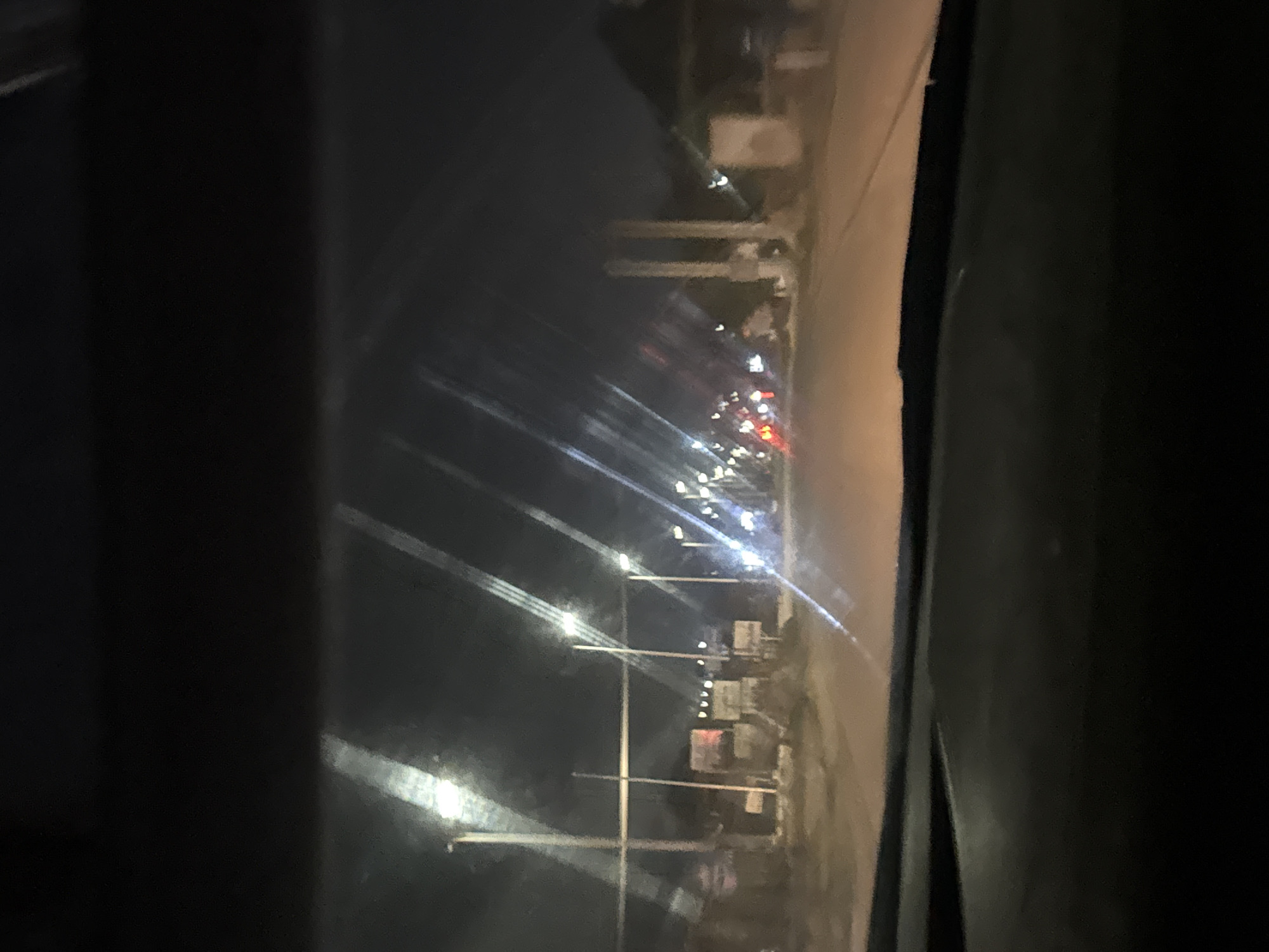 An image, captured at night from the interior of a vehicle, displays a street scene in Gura Top, Nigeria. The perspective is through a windshield, featuring notable reflections and streaks of light across its surface.
The central subject is a road illuminated by numerous artificial lights. Bright white streetlights appear as prominent, elongated streaks across the frame, suggesting either motion blur or lens flare. Further down the road, red and white dots indicate the taillights and headlights of other vehicles. Along the side of the road, vertical poles, likely for streetlights or utility lines, are visible, extending into the distance. Behind these poles, indistinct shapes suggest buildings or roadside structures.
The dark areas bordering the left and right edges of the primary view are parts of the vehicle's interior, such as a door frame or dashboard, framing the external scene. The overall activity depicted is being a passenger or driver in a vehicle traveling along a lit road at night. The time of day is clearly night, evidenced by the pervasive darkness and reliance on artificial illumination. No discernible text is visible anywhere in the image or its crops. The scene conveys the atmospheric glow of urban or semi-urban street lighting during a nighttime journey.