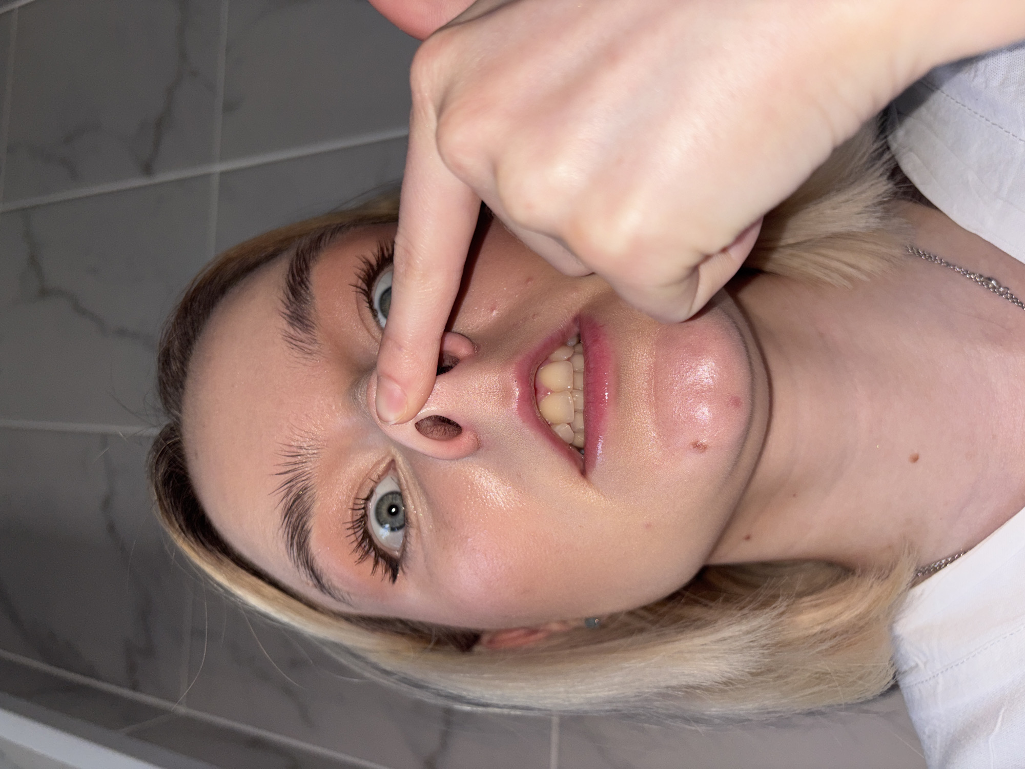 A rotated, close-up portrait captures a person, likely female, with blonde hair and light-colored eyes, in what appears to be an indoor setting with tiled walls. Her head is tilted to the side, and her right hand is prominent in the foreground.
The person's right index finger is pressed against the tip of her nose, pushing it upwards, creating a somewhat playful or humorous expression. Her mouth is slightly agape, revealing both upper and lower teeth, which appear a natural off-white tone. She has long, dark eyelashes and well-defined eyebrows. Her skin exhibits a natural sheen, with a few small moles or blemishes visible on her cheek and neck area. She is wearing a light-colored garment, possibly a white shirt, and a thin silver chain necklace is visible around her neck.
The background consists of light grey tiled walls with subtle marbling patterns and horizontal grout lines, characteristic of an indoor space like a bathroom or kitchen. The lighting is soft and even, highlighting her facial features. There is no visible text in the image. The time of day and external weather conditions cannot be determined from this indoor shot. While the location context is Ladozhskoe Ozero, Russia, no specific visual cues within the image confirm this geographical detail.