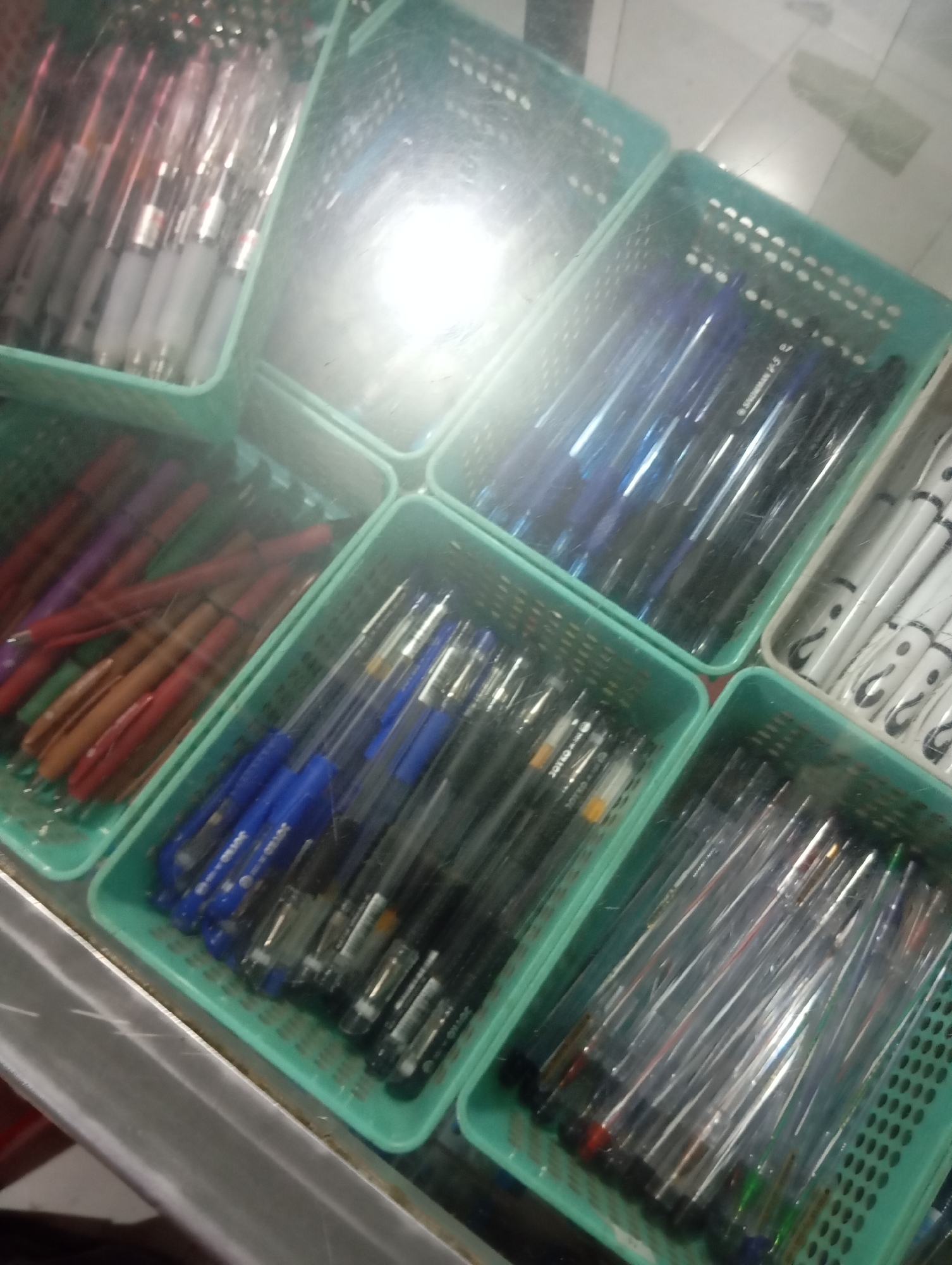 The image shows a top-down view of several teal plastic baskets filled with pens. The pens are organized by color and type. The baskets are arranged in a grid, with some overlapping. The lighting in the image is bright and diffused, possibly from overhead fluorescent lights. The surface beneath the baskets appears to be a counter or table. This scene is located in a stationery shop or a similar retail environment in Sliyeg, Indonesia.