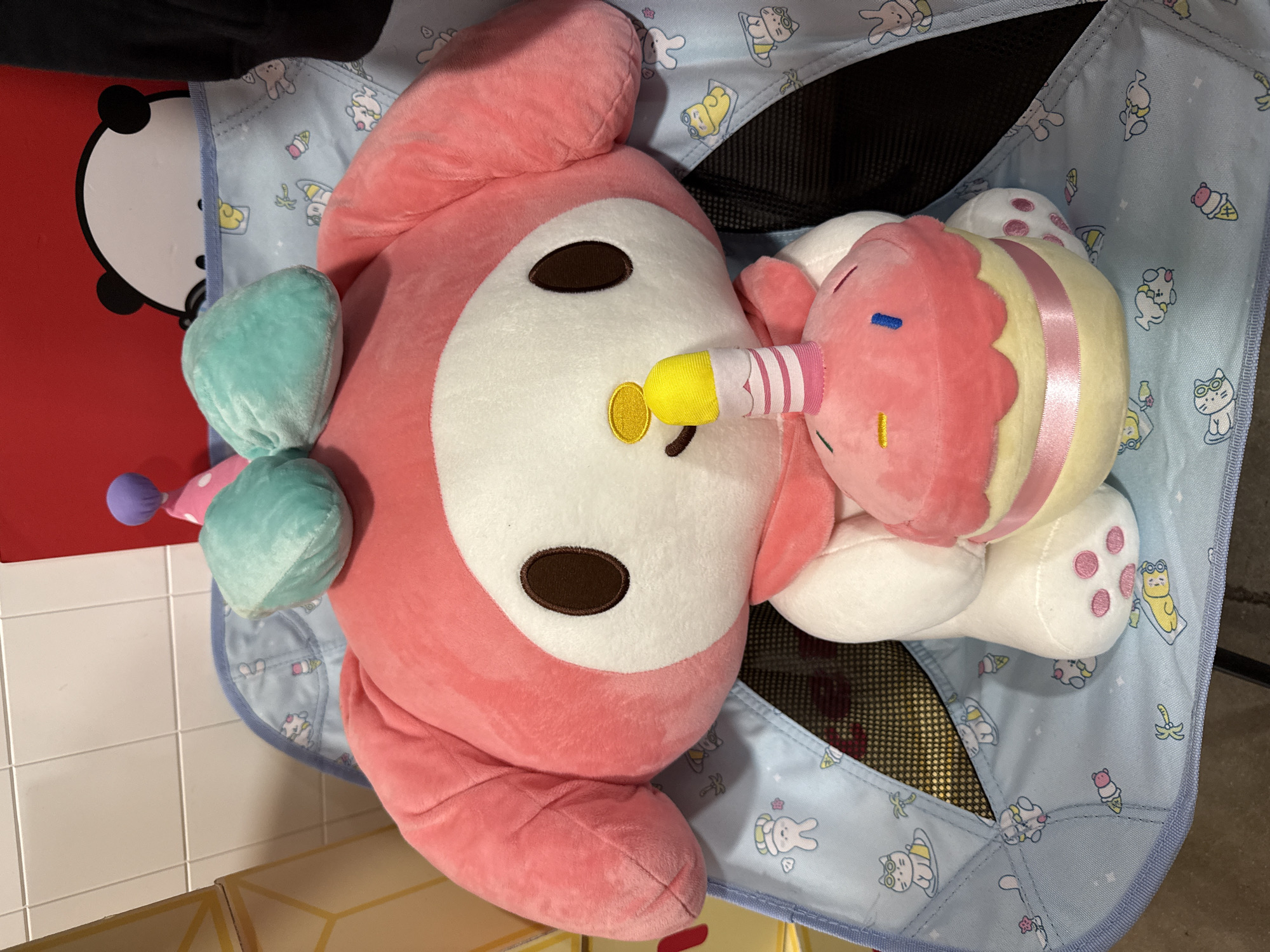 The image shows a close-up shot of two plush toys inside a light blue, patterned child's chair. The larger toy is predominantly pink with a white face and brown eyes, and a yellow object seemingly protruding from its mouth. The second, smaller toy is shaped like a cupcake, with pink and yellow layers, and a pink and white striped "candle." The chair has a light blue background with cartoon images of various animals and objects such as rabbits, cats, ice cream cones, and bananas. The mesh lining of the chair is visible beneath the toys. A red object with a white panda face is partially visible on the left side. The image was taken indoors, possibly in a retail setting. There is no indication of the specific location within Orlando, United States. No people are visible.