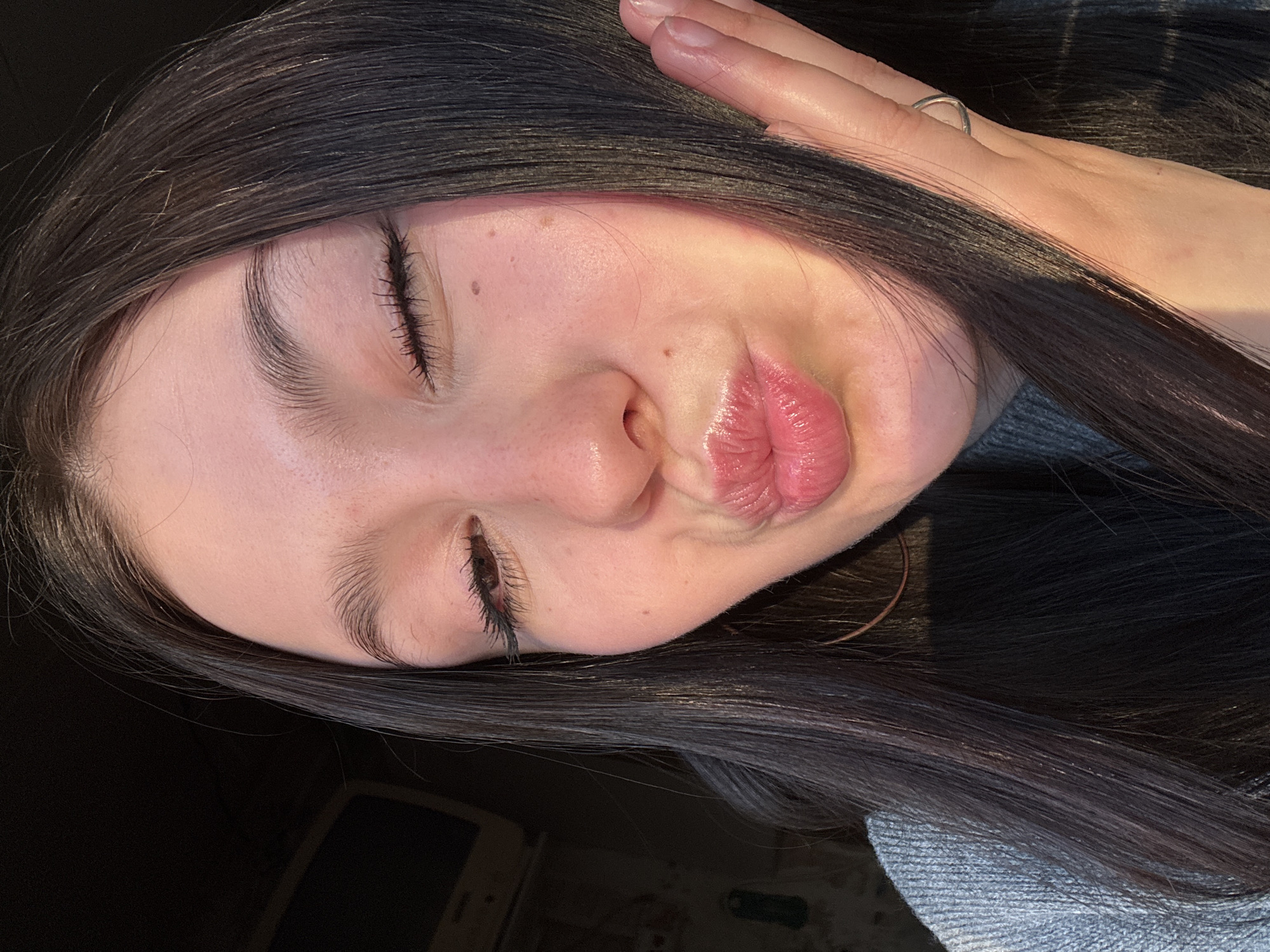 A close-up, rotated image shows a person with dark hair and closed eyes pouting. The person's right hand is near their head. The person is wearing a ring on their hand. The environment is dark, and a portion of a car is visible in the lower left corner. The location cannot be determined from the image.