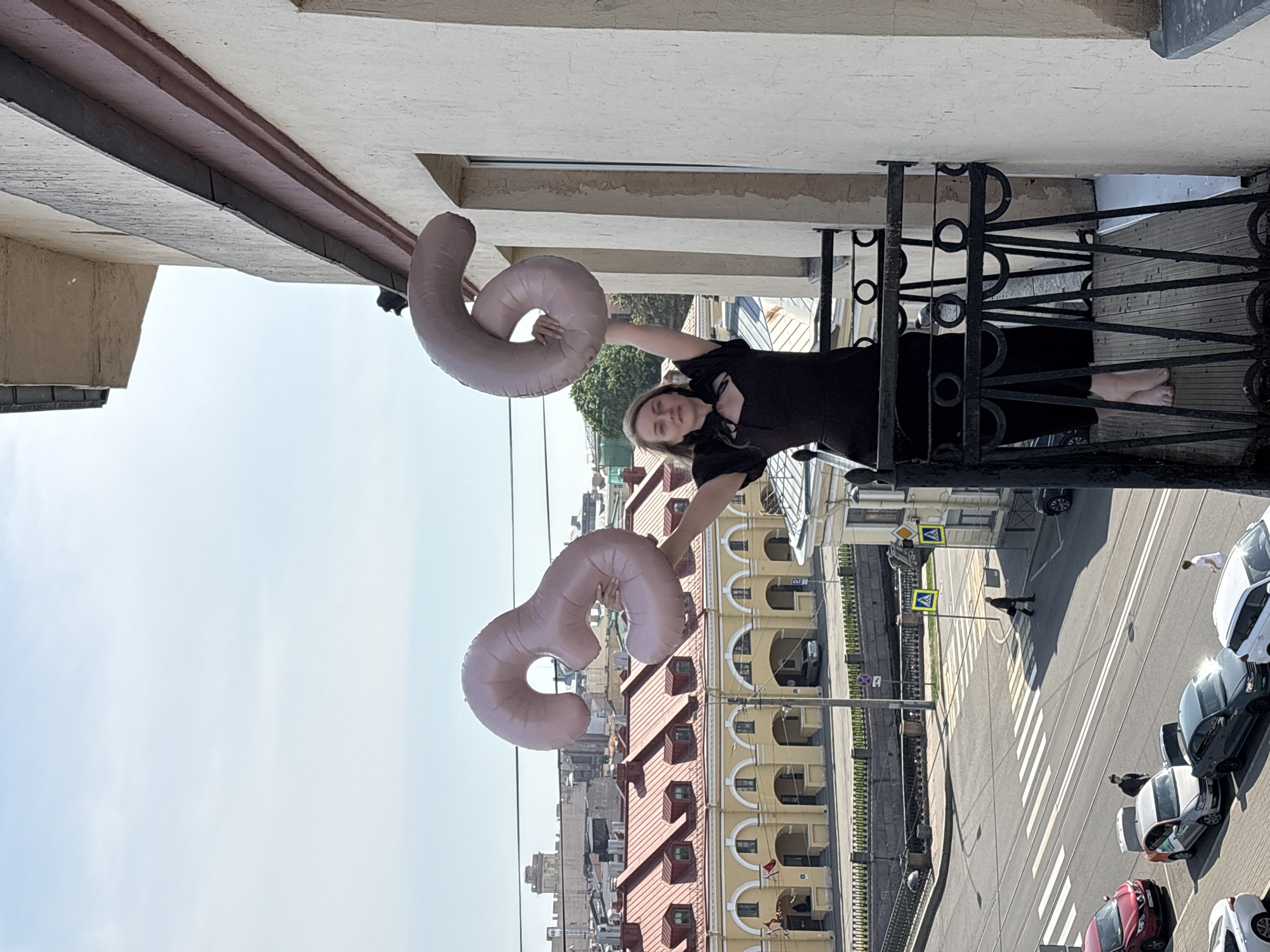The image is taken from a high vantage point, likely a balcony in Saint Petersburg, Russia. The scene depicts a young woman holding two large, inflated number balloons, shaped like a "3" and a "6". She is positioned on a balcony with black metal railings.
Below the balcony, a street scene is visible, featuring a crosswalk and several parked cars. Pedestrians are seen crossing the street. The architecture includes a distinctive yellow building with red roofing and arched window designs.
The sky is clear and bright, suggesting a sunny day. The overall setting is urban, capturing a typical street scene in a Russian city.