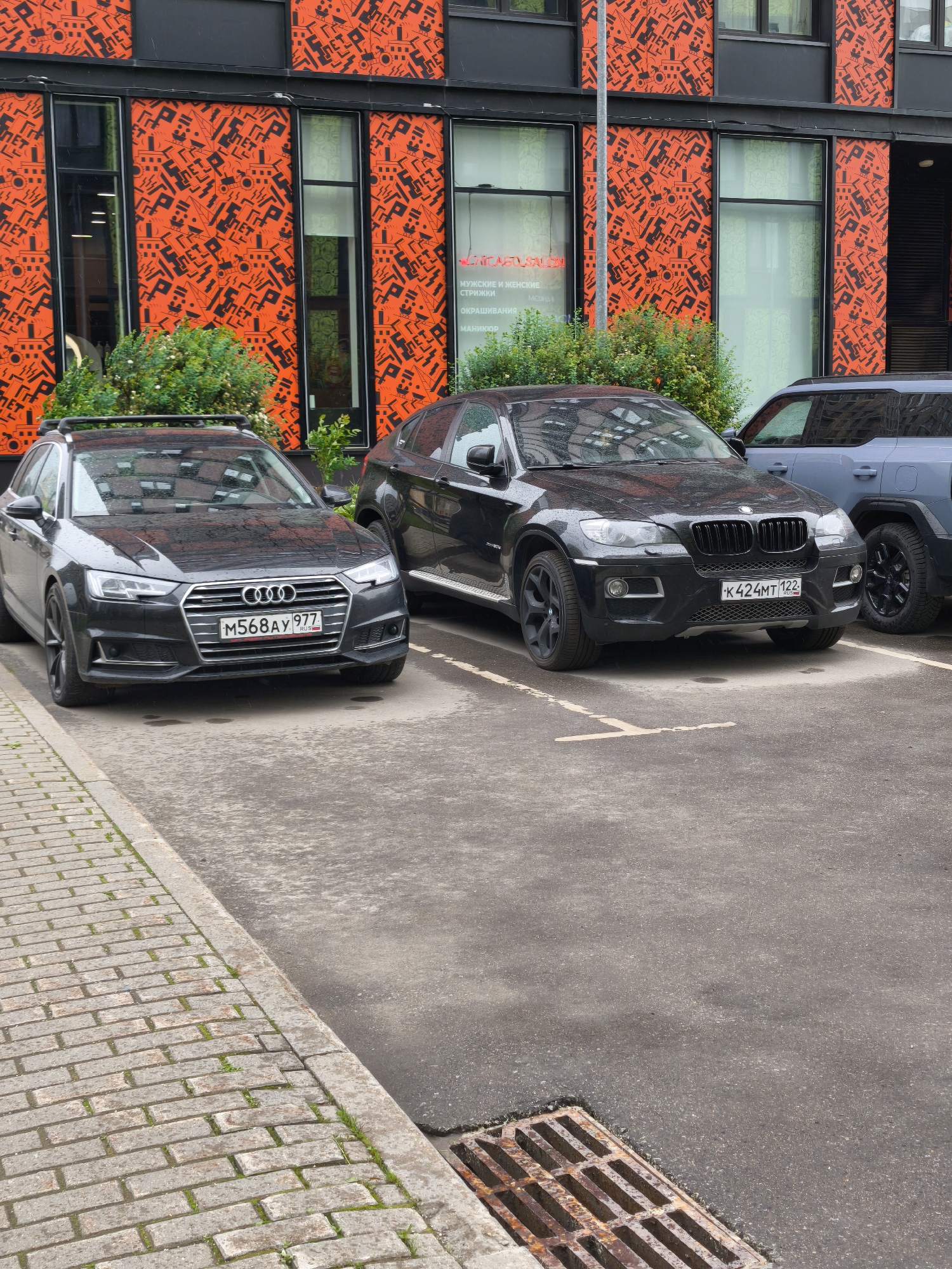 The image shows three cars parked in front of a building with an orange and black patterned facade in Moscow, Russia. The first car on the left is a black Audi A4 Avant with the license plate M568AY 977. Next to it is a black BMW X6 with the license plate K424MT 122. The third car, parked behind the BMW, is a gray Jeep Compass.
The building behind the cars has large windows with a distinctive orange background and black geometric patterns. There is a sign on one of the windows advertising men's and women's haircuts, coloring, and manicures.
The parking area is paved with gray asphalt, and a brick sidewalk runs along the left edge of the image. A metal drain grate is visible in the bottom right corner. The weather appears to be overcast or rainy, as the cars are wet.
