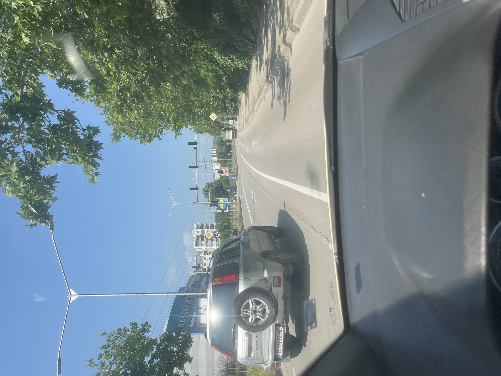 The image captures a sunny daytime street scene from the perspective of a vehicle, looking forward through the windshield. The overall setting is an urban or suburban area in Varna, Bulgaria, characterized by green trees, a clear blue sky, and infrastructure.
In the foreground, the dark interior of the vehicle from which the photo is taken is visible on the right, showing parts of the dashboard or door frame with some reflections. Directly ahead, a silver Honda CR-V SUV is stopped, likely in traffic. It has a spare tire mounted on its rear door and a Bulgarian license plate reading "B 9192 HC." The "B" prefix indicates a vehicle registered in Sofia, Bulgaria.
The road ahead is asphalt, with white lane markings. To the left, there's a curb lined with lush green trees casting shadows. Further down the road, multiple traffic lights are visible, and beyond them, residential buildings and other structures. A prominent yellow and white diamond-shaped "priority road" sign is visible on the left side of the road. Close to the traffic lights, a blue circular "no U-turn" sign can be seen.
On the left side, a large sign or building facade displays the text "RESERVED" and partially "House DECOR". The sky is clear blue, indicating good weather. The bright sunlight suggests it is mid-day or early afternoon.