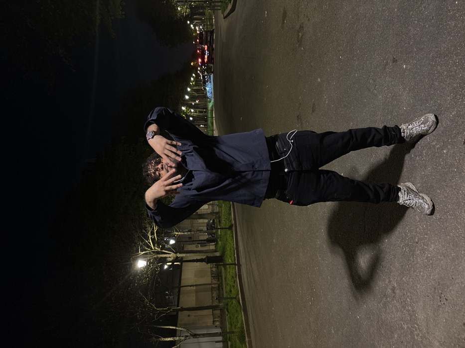 A single male individual stands centered on a dark, paved urban road at night. He is facing the camera, with both hands raised to chest height, palms outward and fingers slightly curled, partially obscuring his lower face. He wears a dark blue long-sleeved collared shirt, black jeans, and silver-toned athletic shoes. A wristwatch is visible on his left wrist, and white wired earbuds are hanging from his right front jean pocket. A distinct, elongated shadow of the subject is cast to his immediate left on the asphalt.
The scene is set on a wide, tree-lined avenue characteristic of Paris, France. Numerous classic Parisian-style lampposts with distinctive globe-shaped lights illuminate the roadway and sidewalks stretching into the background on both sides. To the left, behind a strip of grass and trees, a light-colored building with a dark, ornate fence is partially visible. To the right, in the distance along the avenue, several vehicles are identifiable by their illuminated headlights and taillights. The dense, dark foliage of the trees against the night sky frames the upper portion of the image.