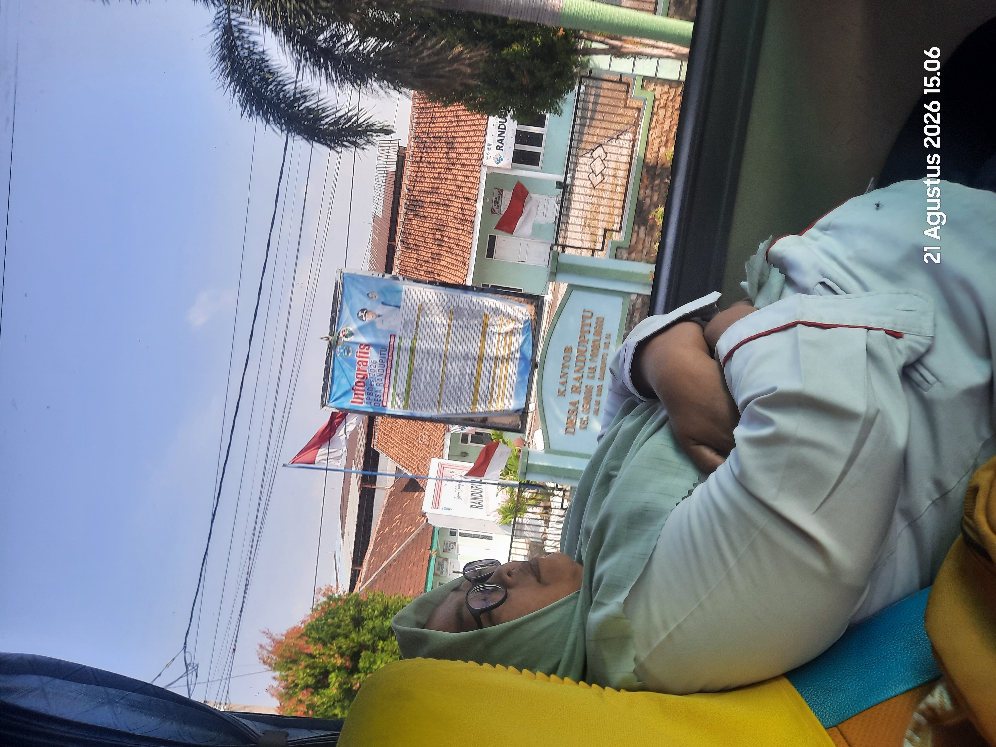 A medium shot shows a woman wearing a pale green hijab and glasses, sleeping on a bus. She is dressed in a white uniform with red trim. The woman is leaning against a yellow headrest. The bus window offers a view of a street scene in Gending, Indonesia. Buildings with red-tiled roofs, a palm tree, and a billboard are visible. Two Indonesian flags are displayed, one on a building and another on a pole. The billboard features an infographic and portraits of two men. The date "21 Agustus 2026 15.06" is visible in the lower right corner.