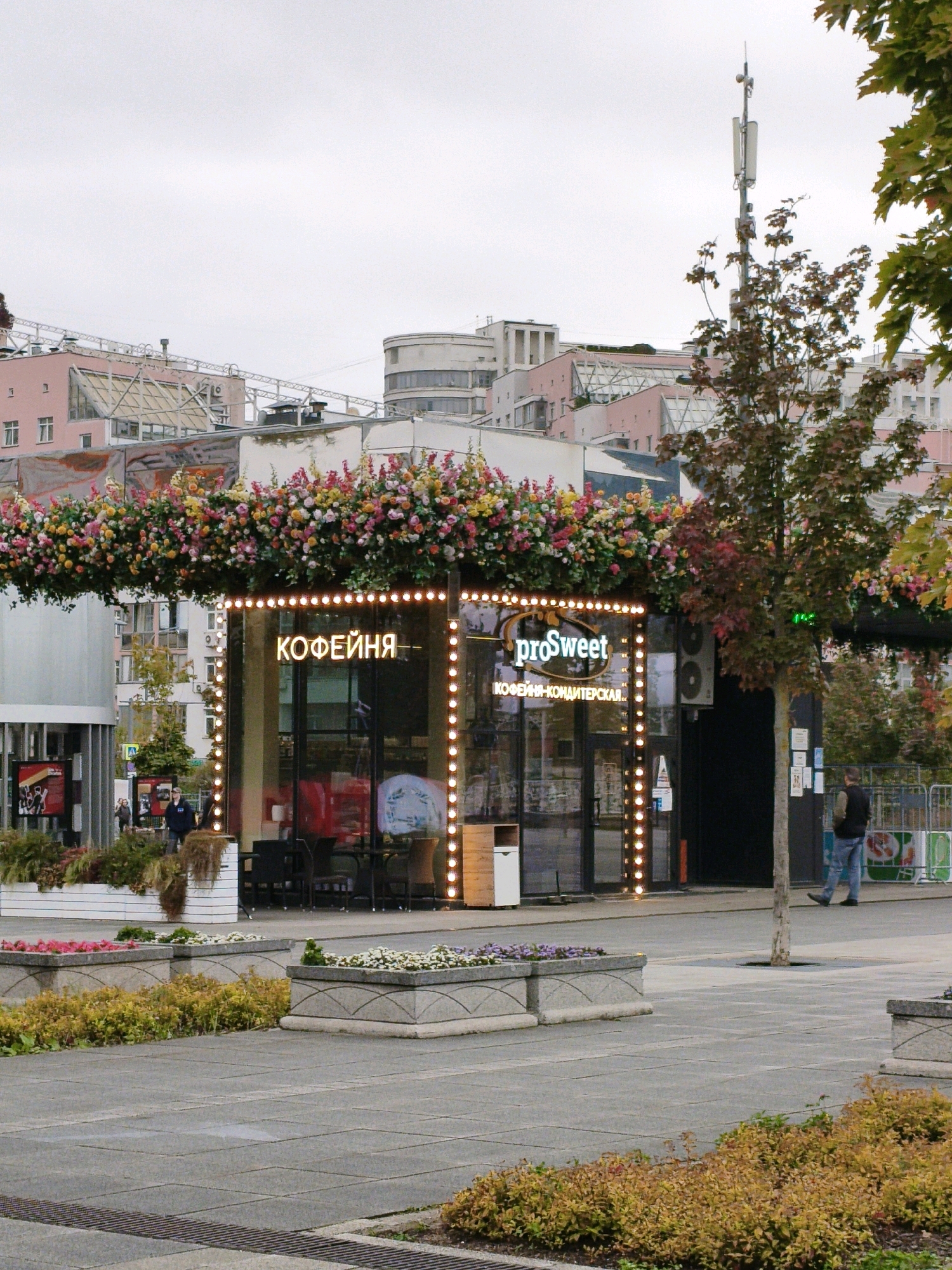 A storefront, identified by the Russian text "КОФЕЙНЯ" (Coffee House) and the logo "proSweet" with the Russian text "КОФЕЙНЯ-КОНДИТЕРСКАЯ" (Coffee House-Confectionery), is adorned with a dense floral arrangement of colorful flowers across its awning. The storefront features large glass windows and is illuminated by a string of lights. Inside, tables and chairs are visible. A person is walking on the paved ground in front of the store, which is lined with planters containing more flowers and greenery. In the background, buildings of various architectural styles are present under a cloudy sky, and a cell tower stands tall. The scene appears to be in an urban setting, possibly a plaza or pedestrian walkway.