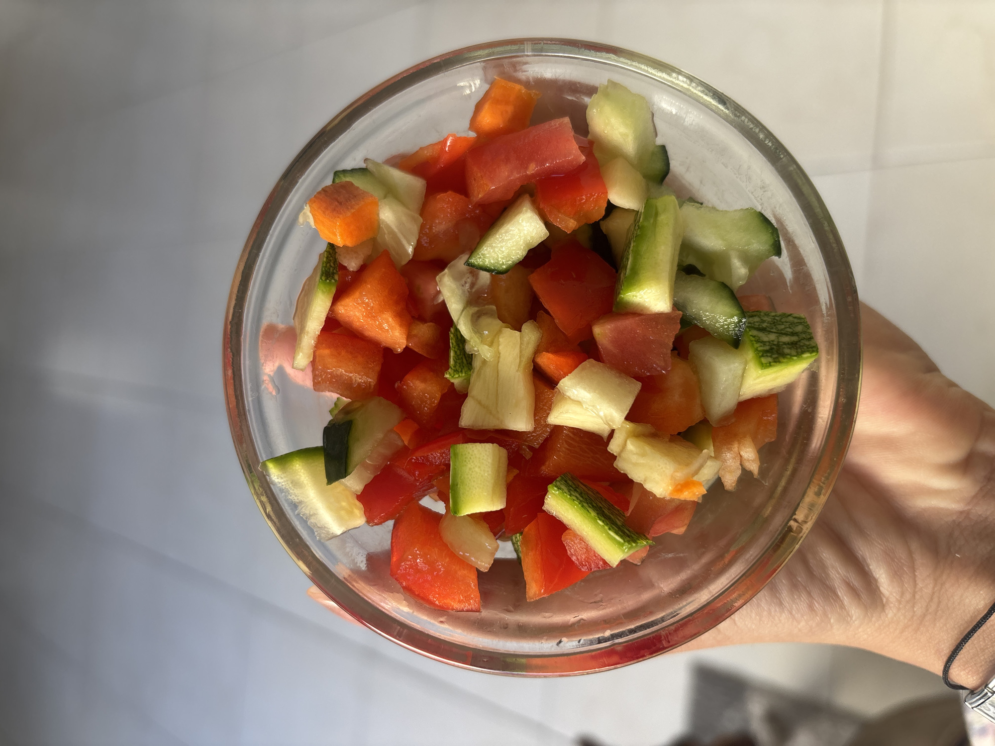 The image shows a glass bowl filled with chopped vegetables. The vegetables appear to include red bell pepper, cucumber, carrot and white squash. The bowl is being held by a person with fair skin, as evidenced by their hand and wrist visible at the bottom right of the image. The background is a plain white or off-white surface.
It is not possible to determine the location as Puebla, Mexico.